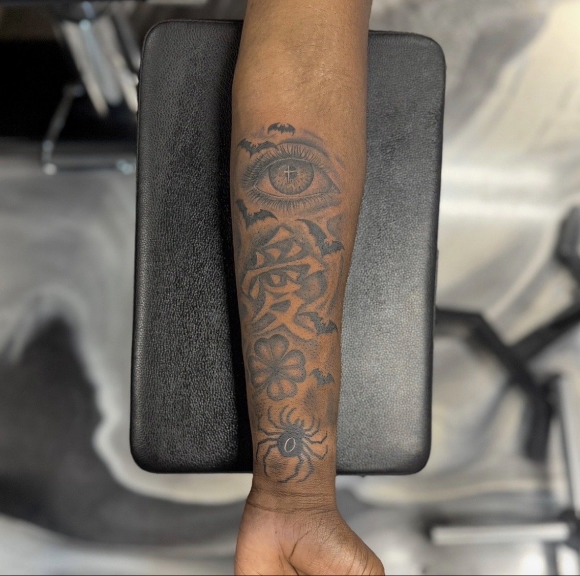 Tattoo on forearm featuring an eye with a cross in the pupil, surrounded by bats, a flower, a spider, and abstract elements.