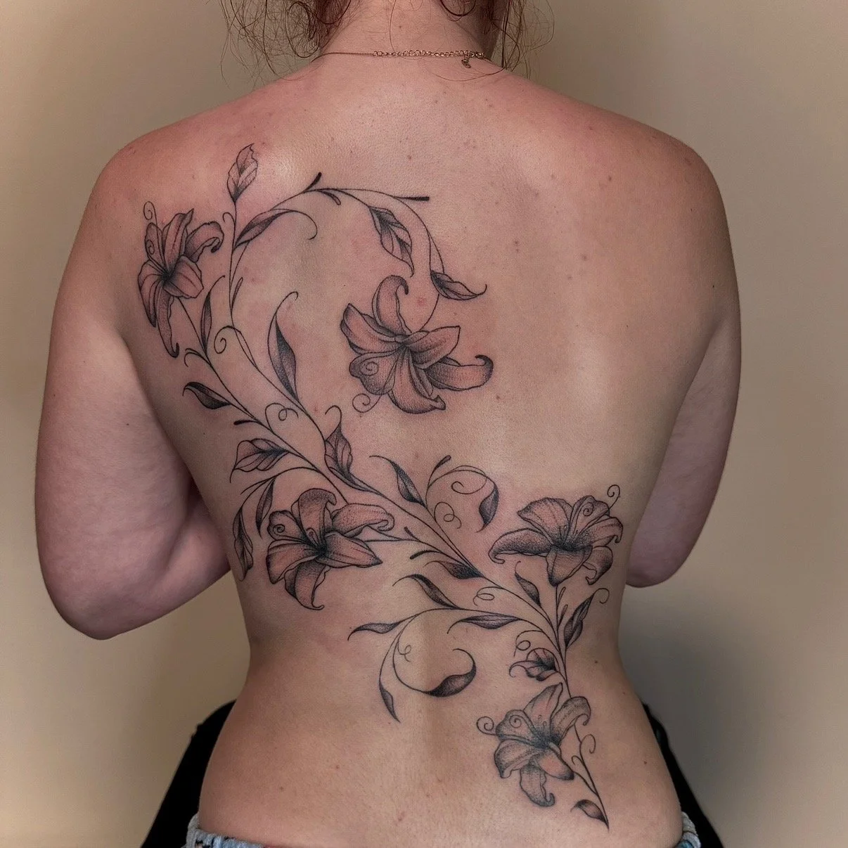 A person's back with a large floral tattoo featuring lilies and leaves in black ink.
