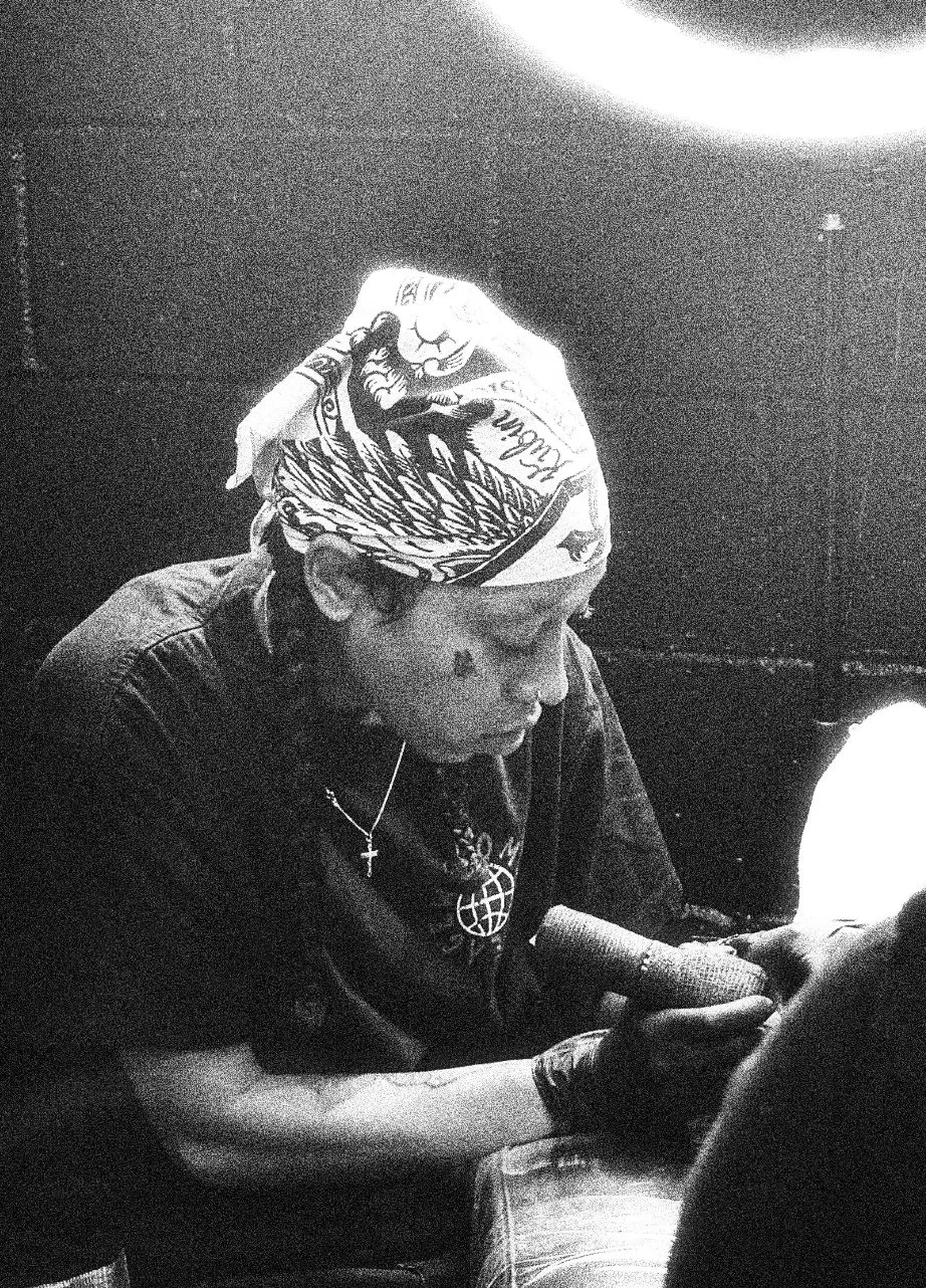 A person with a bandana on their head, wearing a necklace and a T-shirt, is looking at and holding a phone in their right hand, sitting at a table with a microphone and a glass of water, in a dimly lit room.
