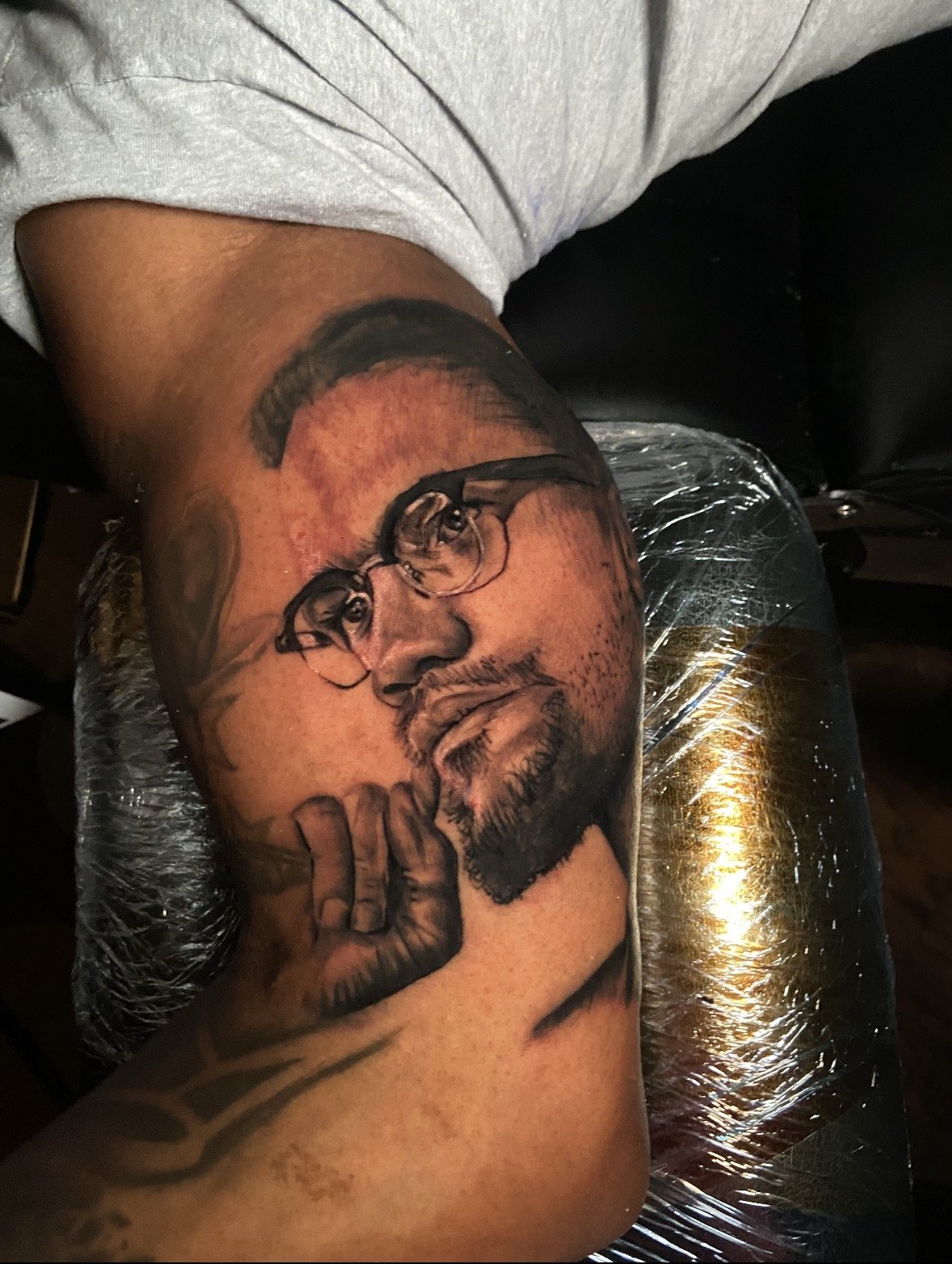 A detailed black and gray portrait tattoo of a man with glasses, a beard, and a finger raised to his lips, on a person's arm.