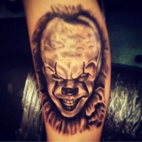 Tattoo of a creepy clown face with a sinister grin, on a person's arm.