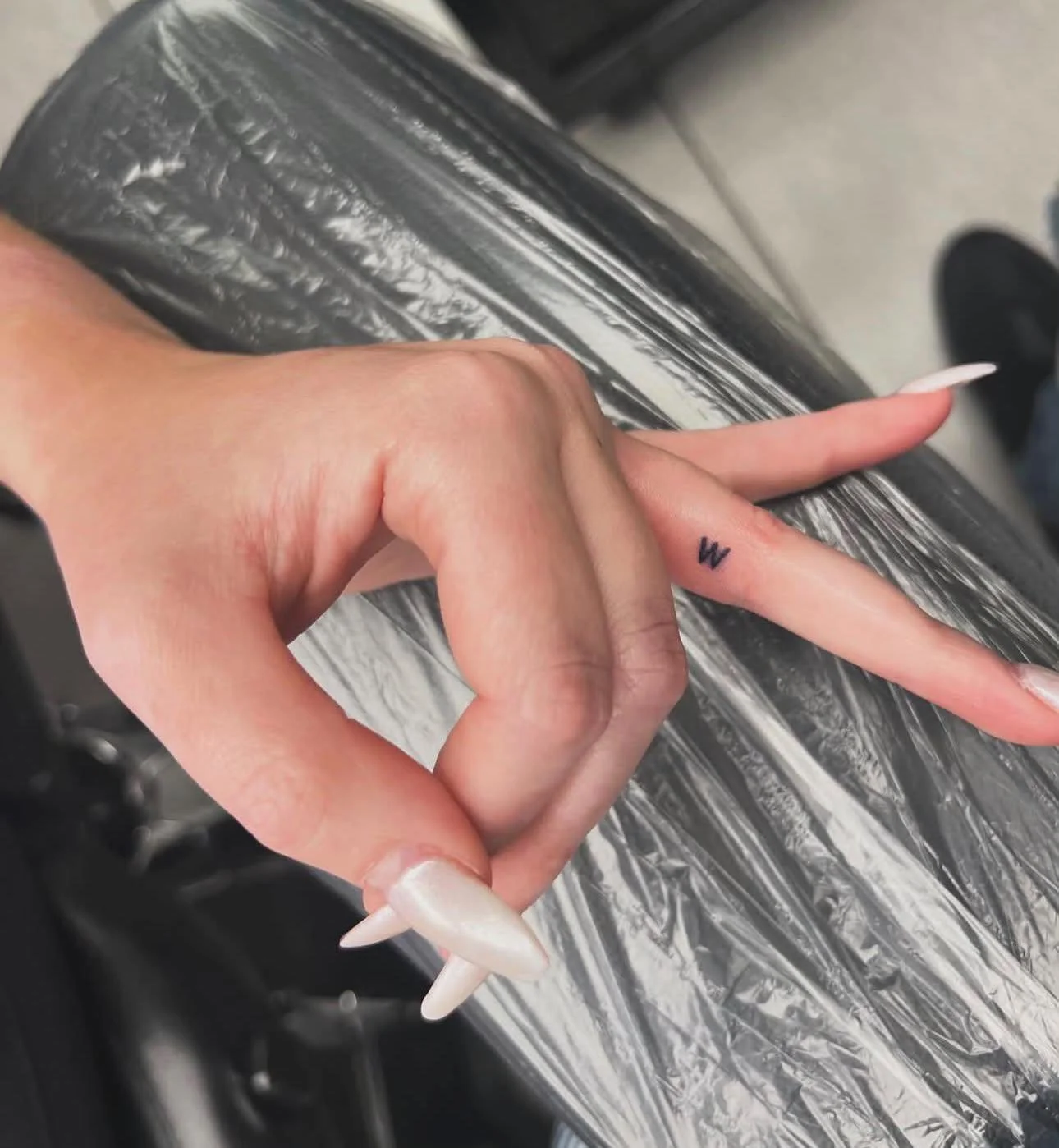 A hand with long, pointed, white nails making a peace sign, resting on a black, shiny, textured surface. The hand has a small, black tattoo of the letter 'W' on the ring finger.