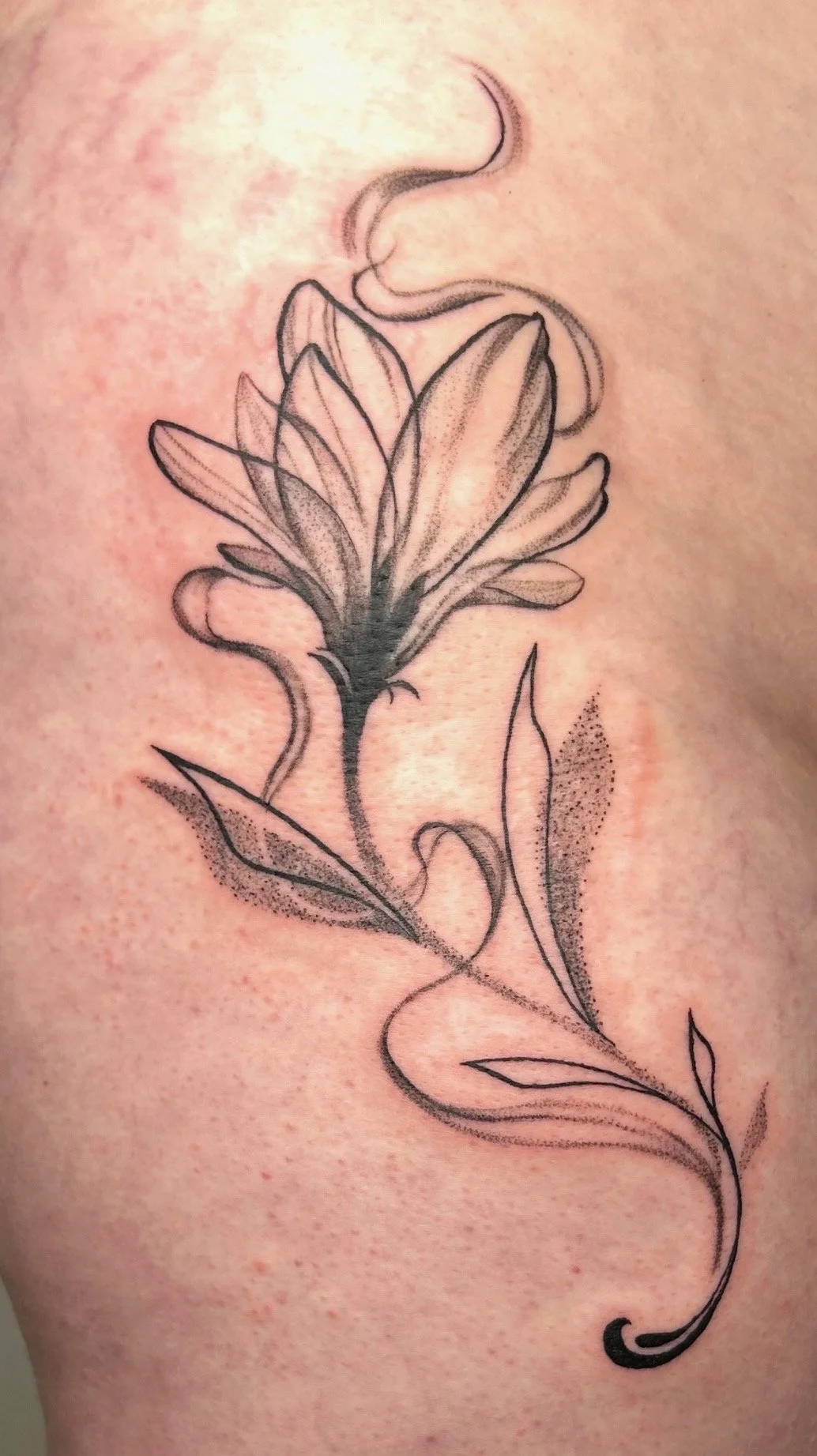 A tattoo of a flower with detailed petals and leaves, rendered in black ink with shading and dot work for depth.