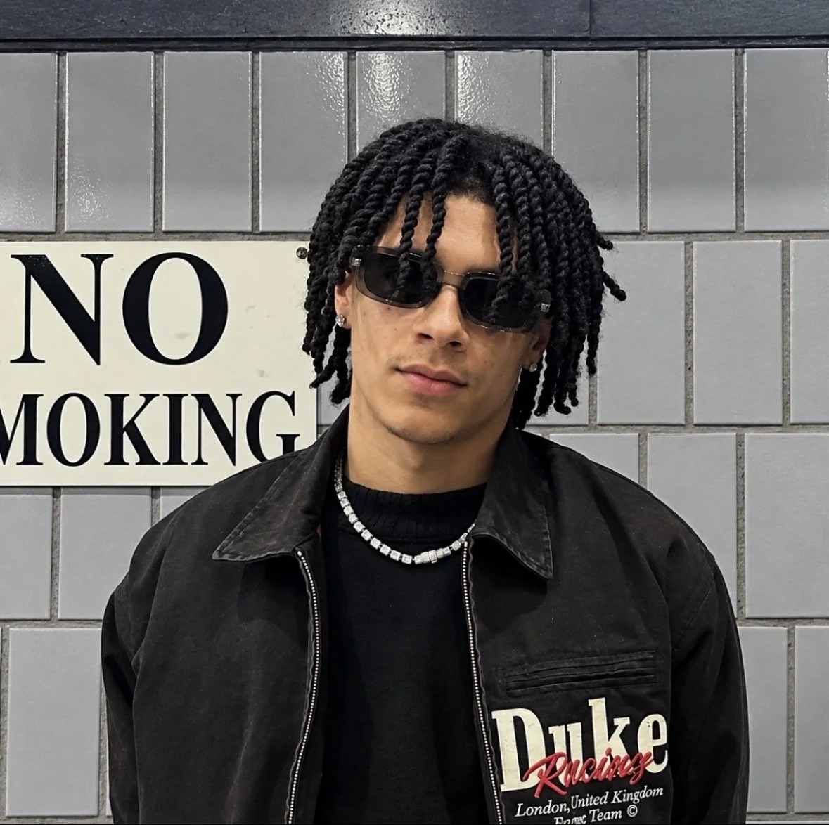 A young man with black twisted hair, sunglasses, and earrings standing in front of a grey tile wall with a 'No Smoking' sign.