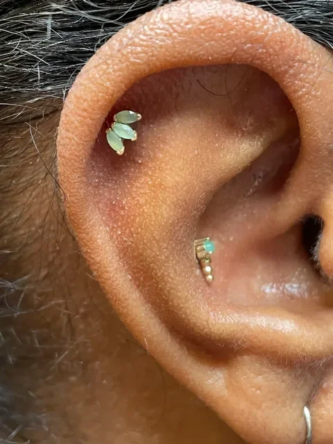 Close-up of a person's ear with two earrings: a cluster of three green gemstone hoops in the upper part and a tiny silver hoop with a turquoise stone in the lower part.