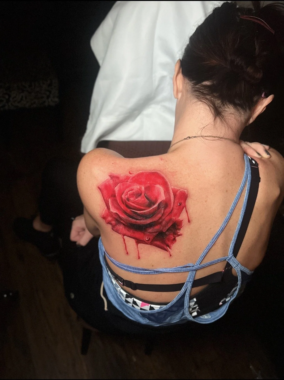 A woman with dark hair in a braid shows a realistic red and black rose tattoo on her right shoulder blade, with shading and dripping ink effects.