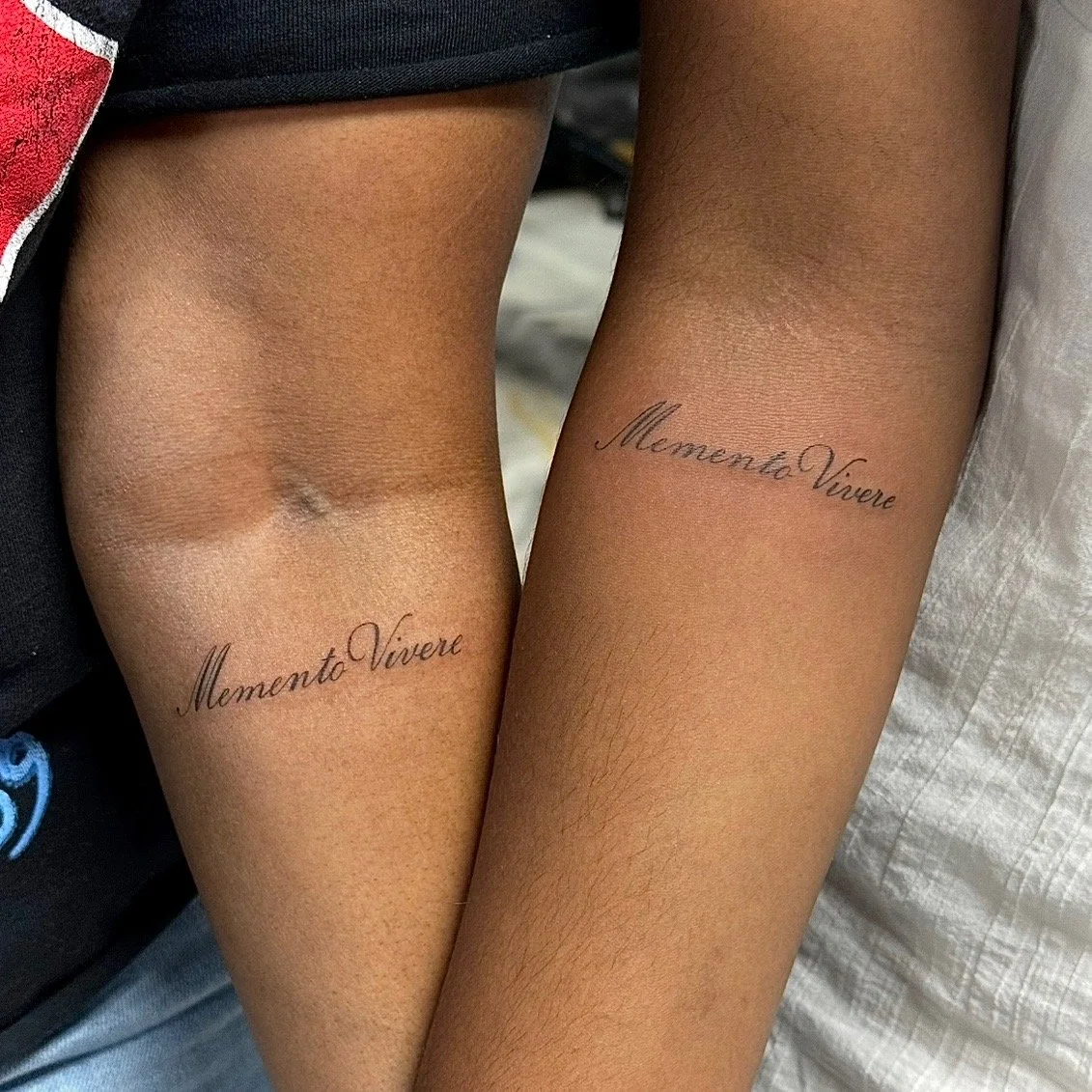 Two forearms with matching tattoos that read 'Memento Vivere' in cursive script.