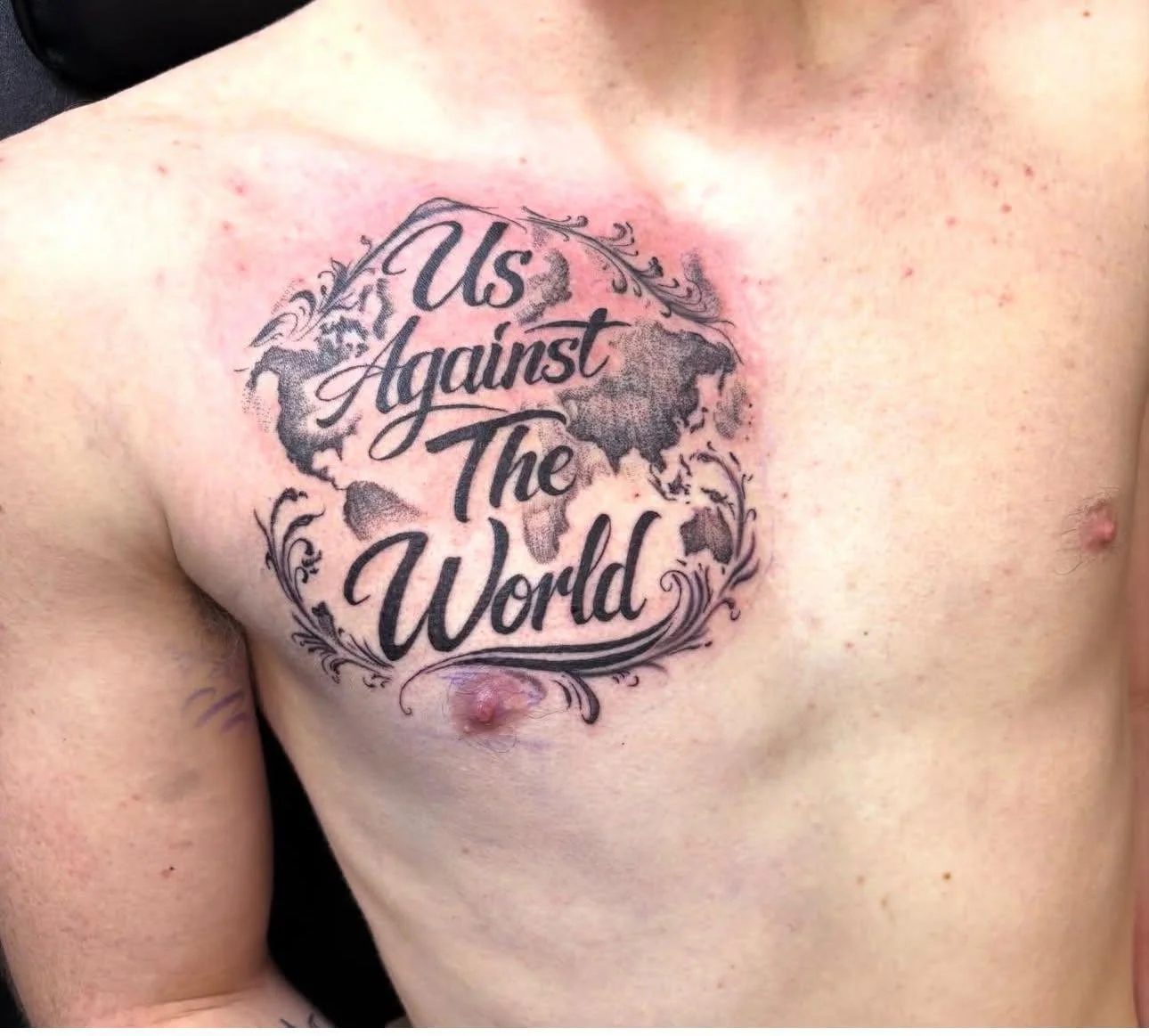 Tattoo on person's chest with a world map and the phrase 'Us Against The World' in cursive lettering.