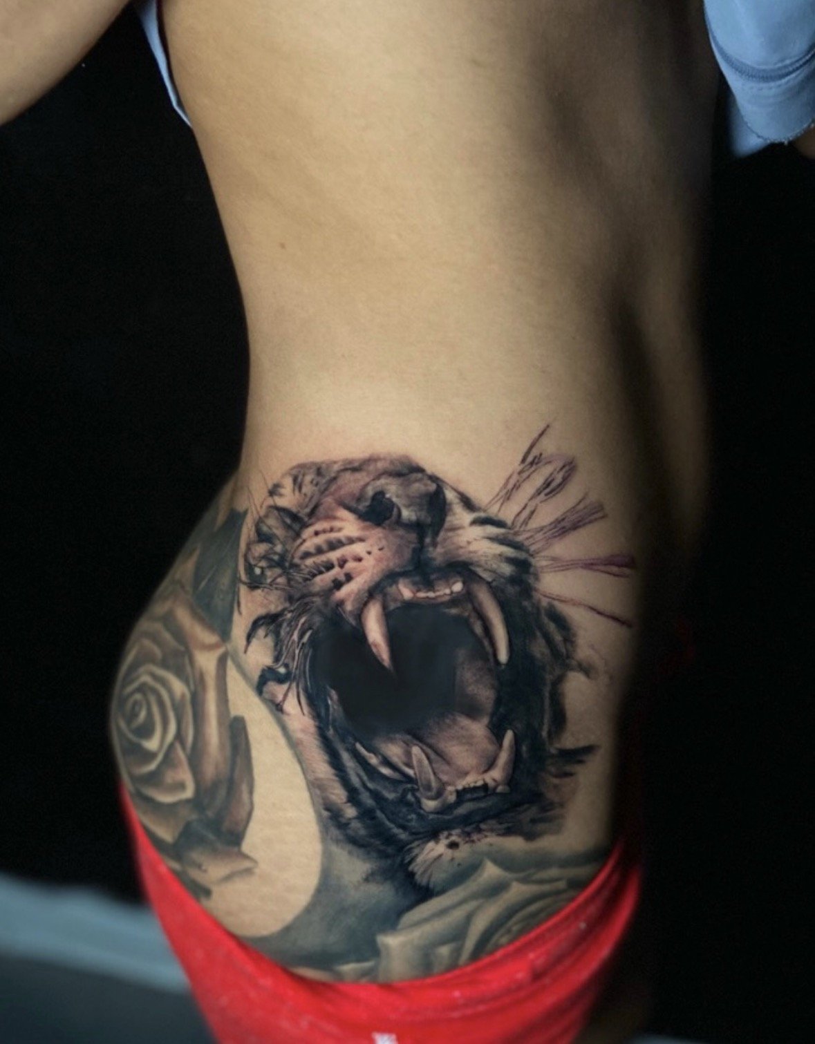Tattoo of a roaring lion's face with open mouth and sharp teeth on a person's side torso.