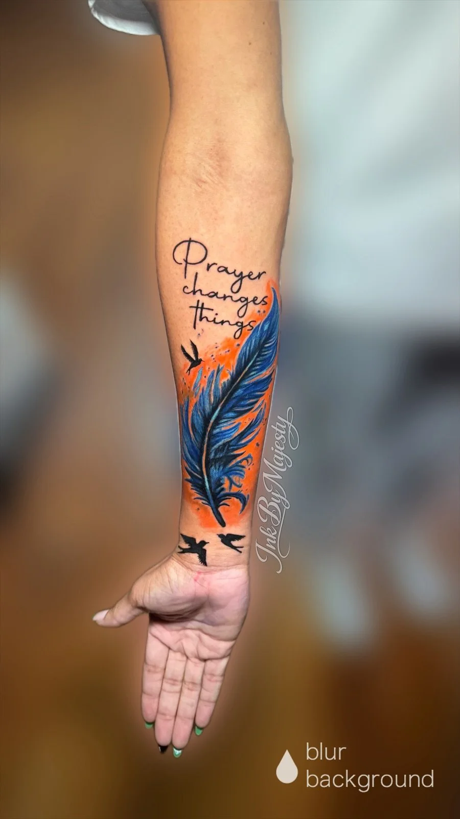 Tattoo on person's forearm featuring a large blue feather with an orange background, accompanied by text that reads 'Prayer changes things' and a small flying bird.