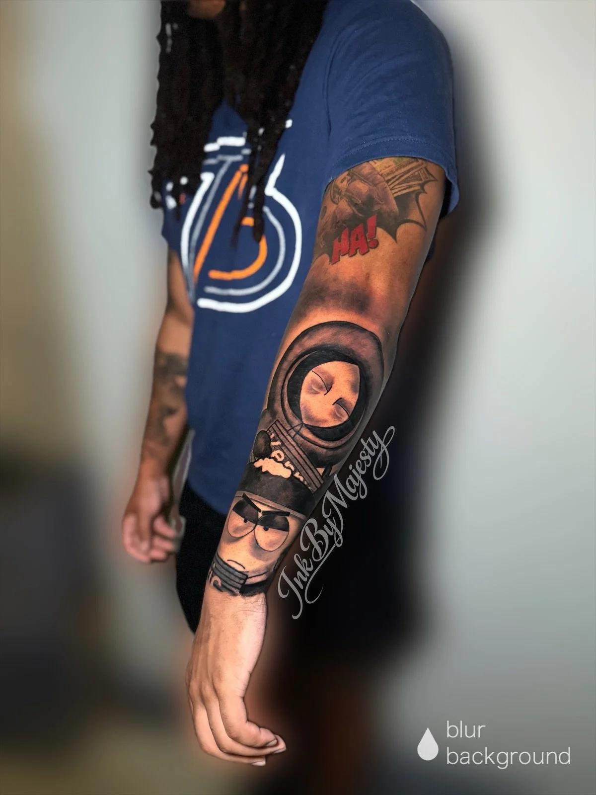 A person's arm with detailed tattoos, including a circular design, a cartoon character with sunglasses, and comic-style text saying "HA!" The person is wearing a blue T-shirt with a white and orange logo, standing against a blurred background.