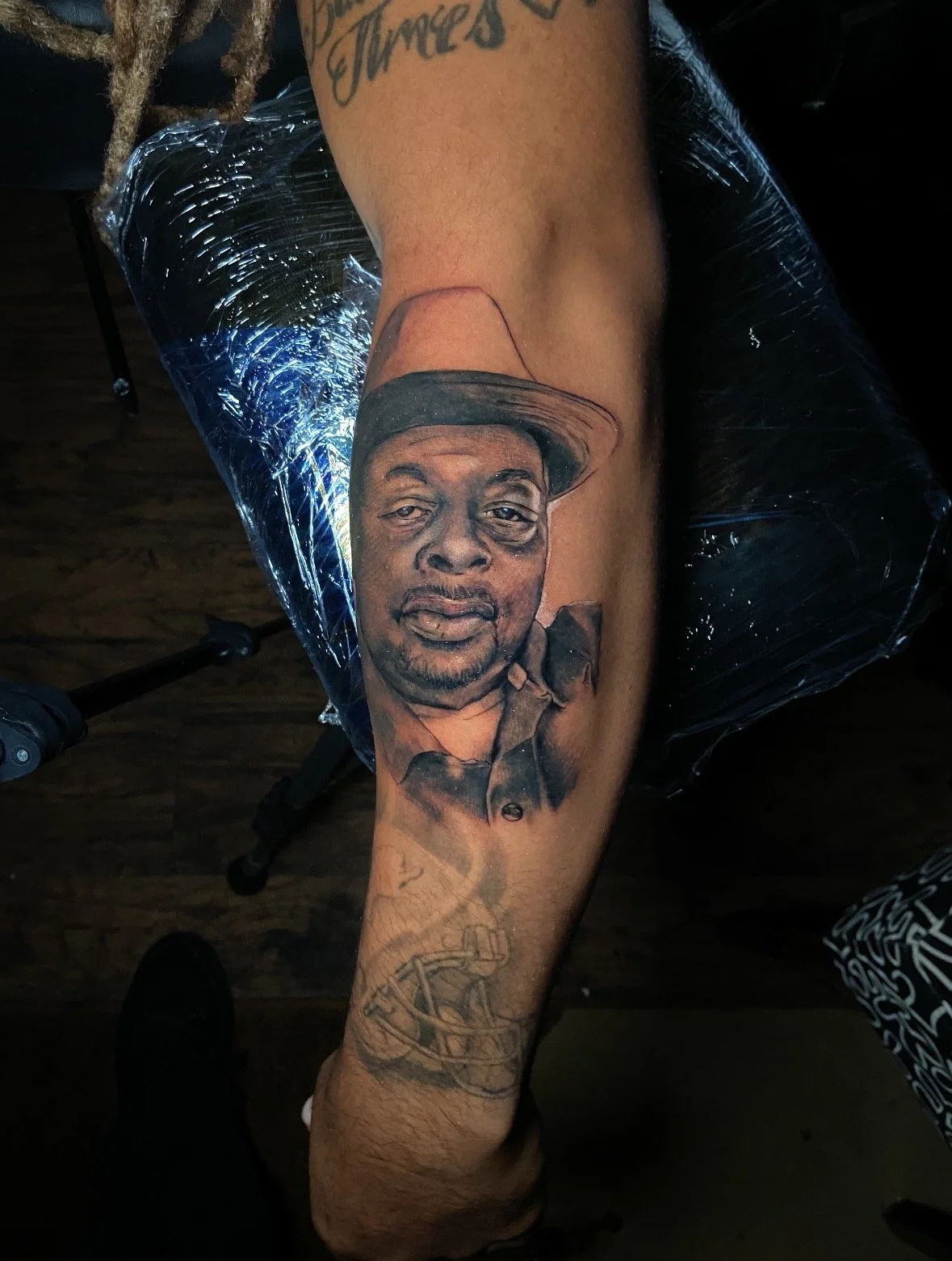 Tattoo of a man's face wearing a hat on someone's arm, with additional tattoos including a compass and a chain.
