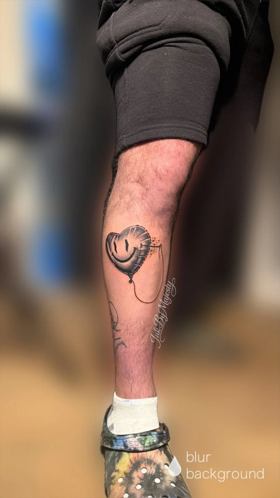 Tattoo of a smiley face balloon with a string tattooed on a person's lower leg, with a blurred background.
