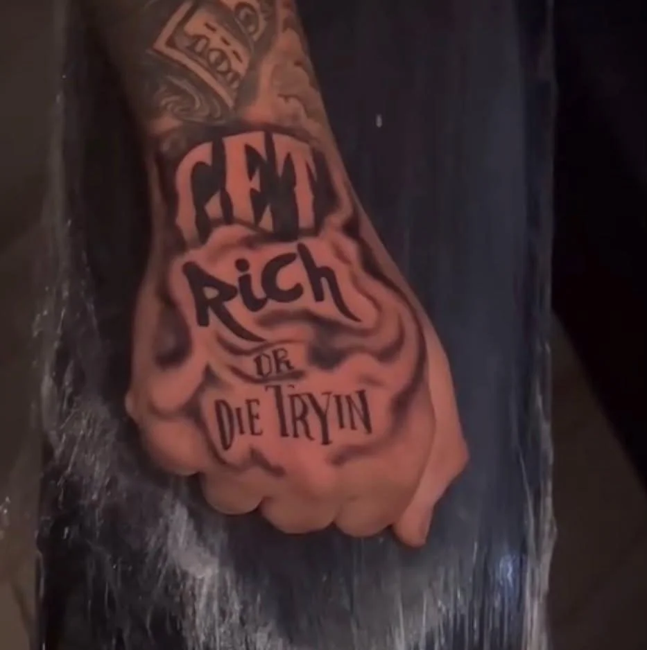 A tattooed arm resting on a surface with a tattoo that reads 'GET RICH OR DIE TRYIN'.