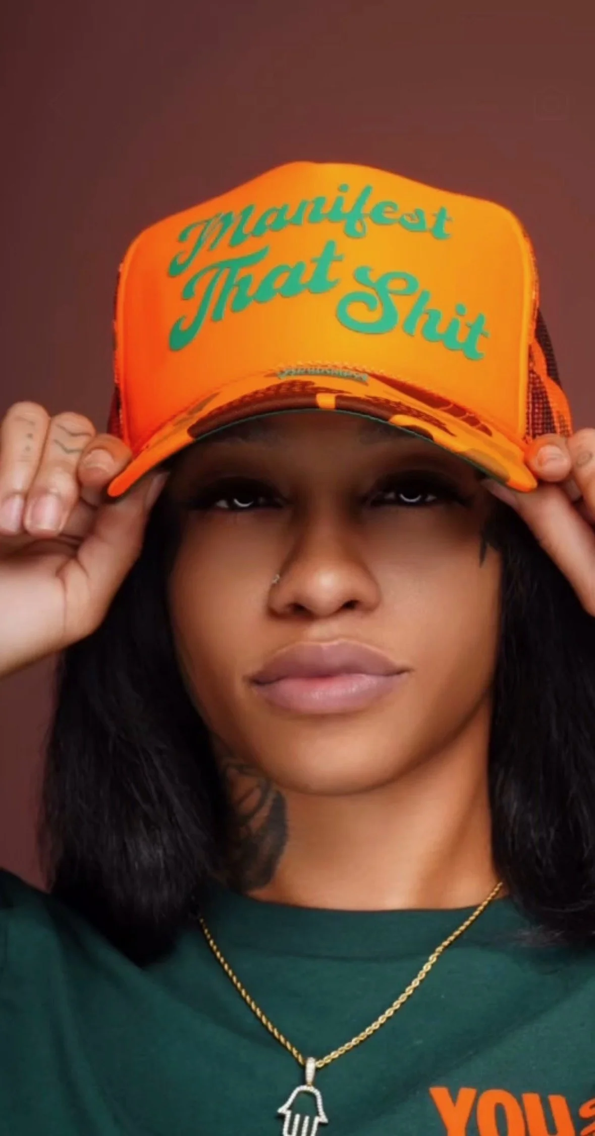 A woman with dark hair and a nose piercing holding an orange trucker hat with teal writing that says "Manifest That Shit".