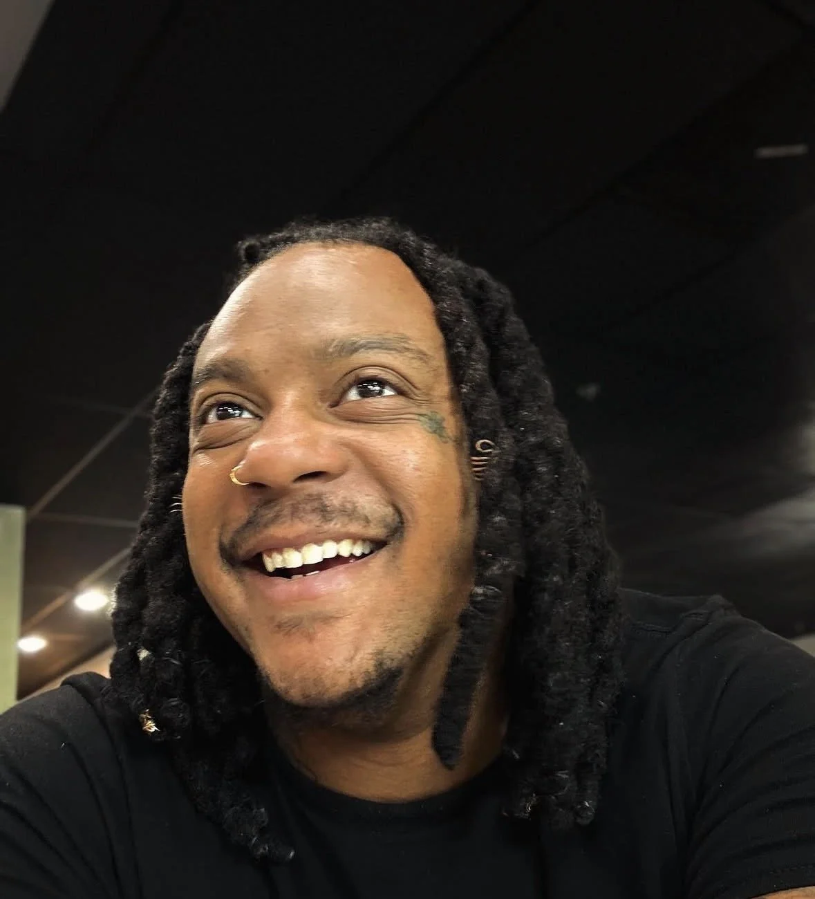Close-up of a smiling man with dreadlocks, wearing a nose ring and earrings, in a dimly lit indoor setting.