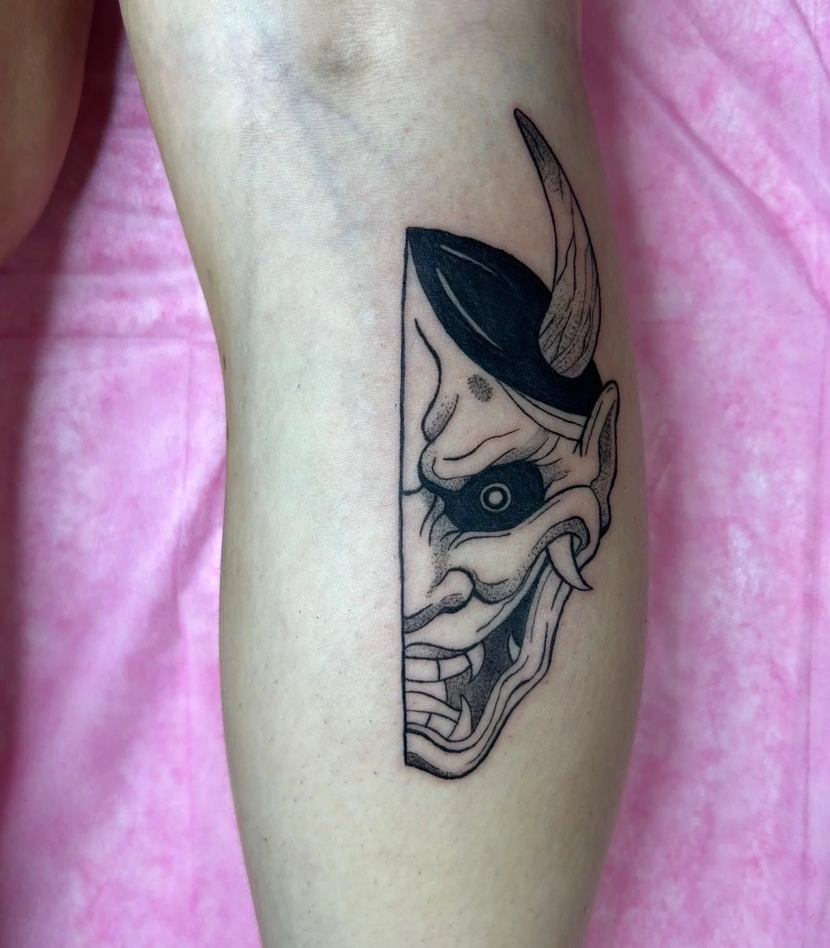 Tattoo of a demon or oni mask on a person's upper arm, with dark black and gray ink, featuring sharp teeth, a hole in the middle, and exaggerated facial features.