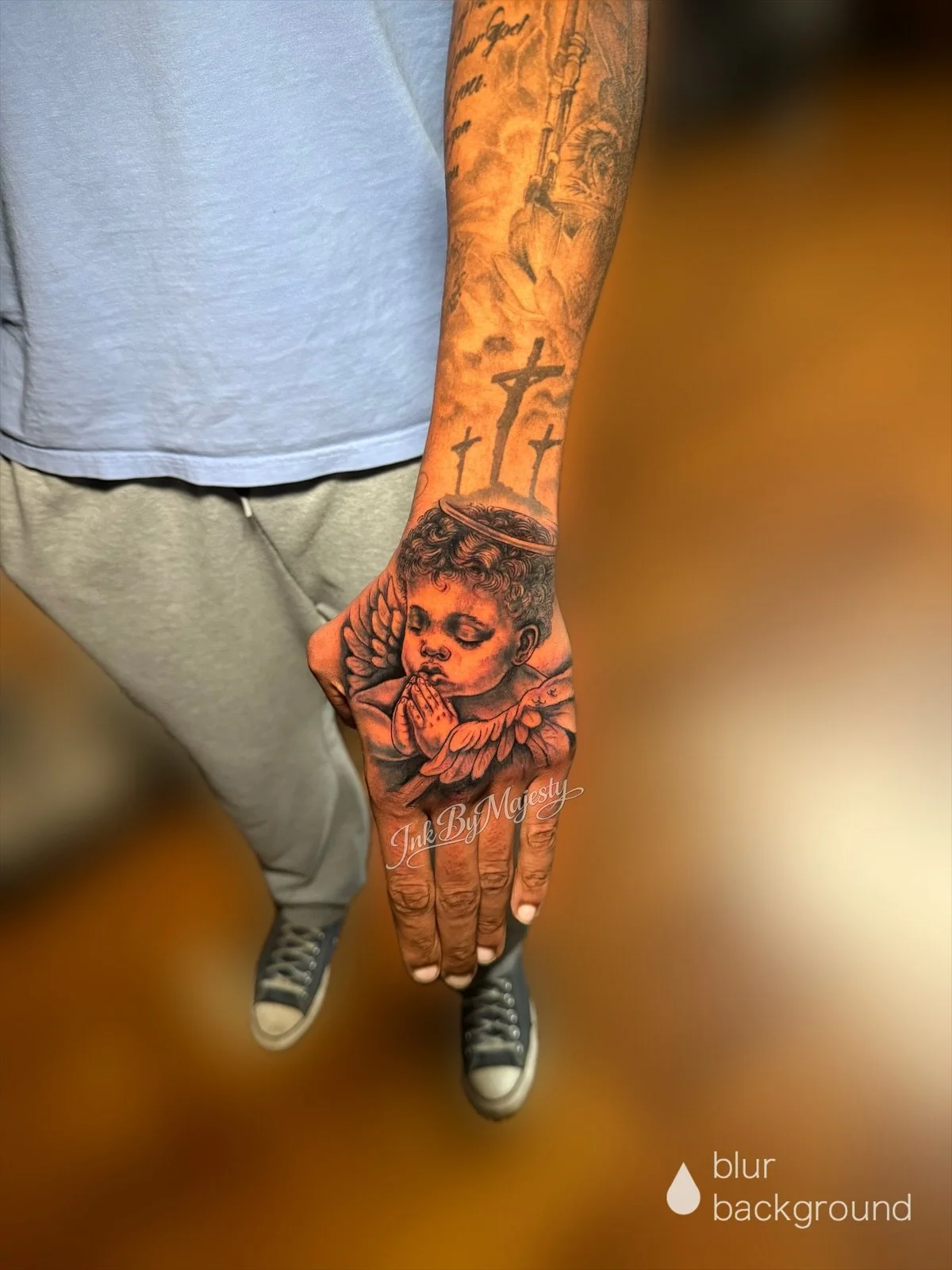 Tattooed arm with a Hindu goddess, praying angel, and swords, wearing a blue shirt and gray pants, standing on a wooden floor.