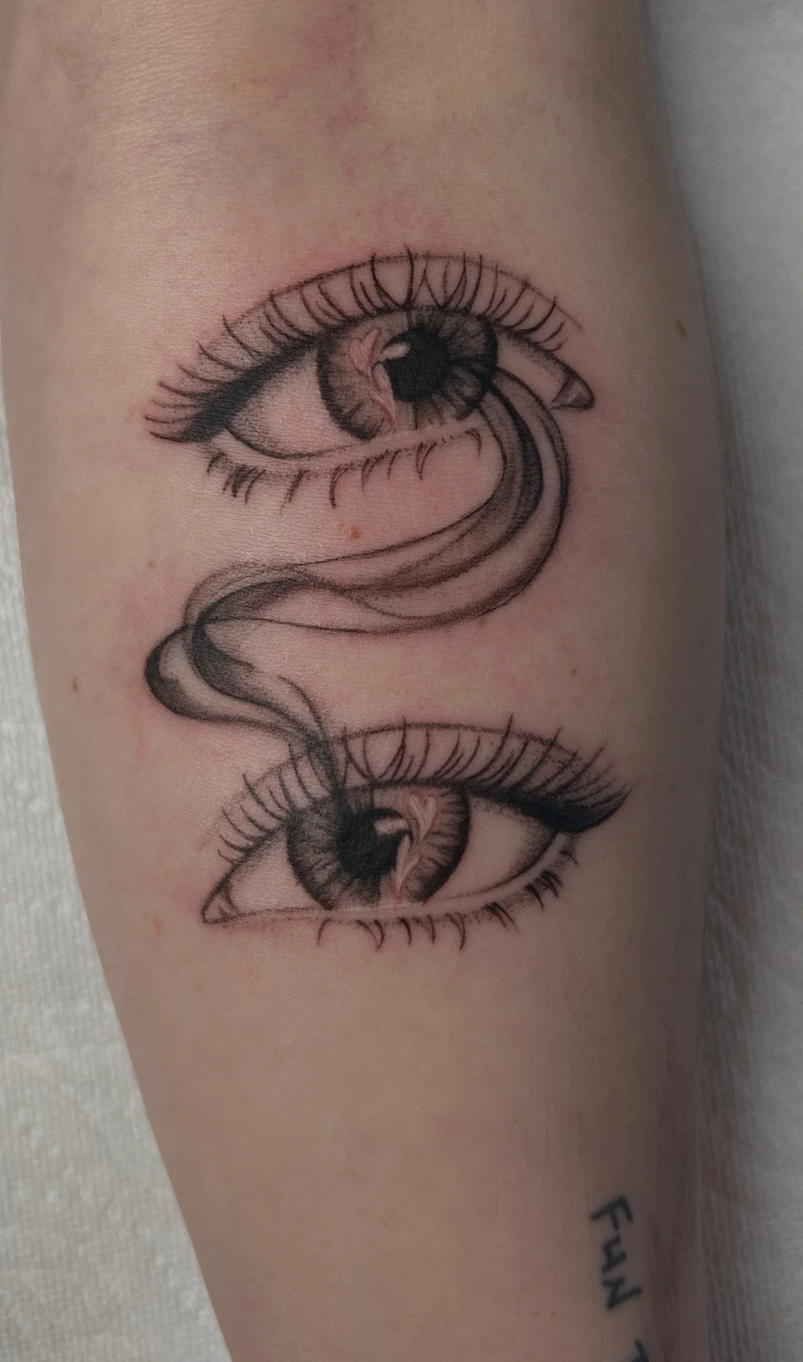 Tattoo of two detailed eyes with eyelashes and irises, connected by a flowing ribbon-like design.