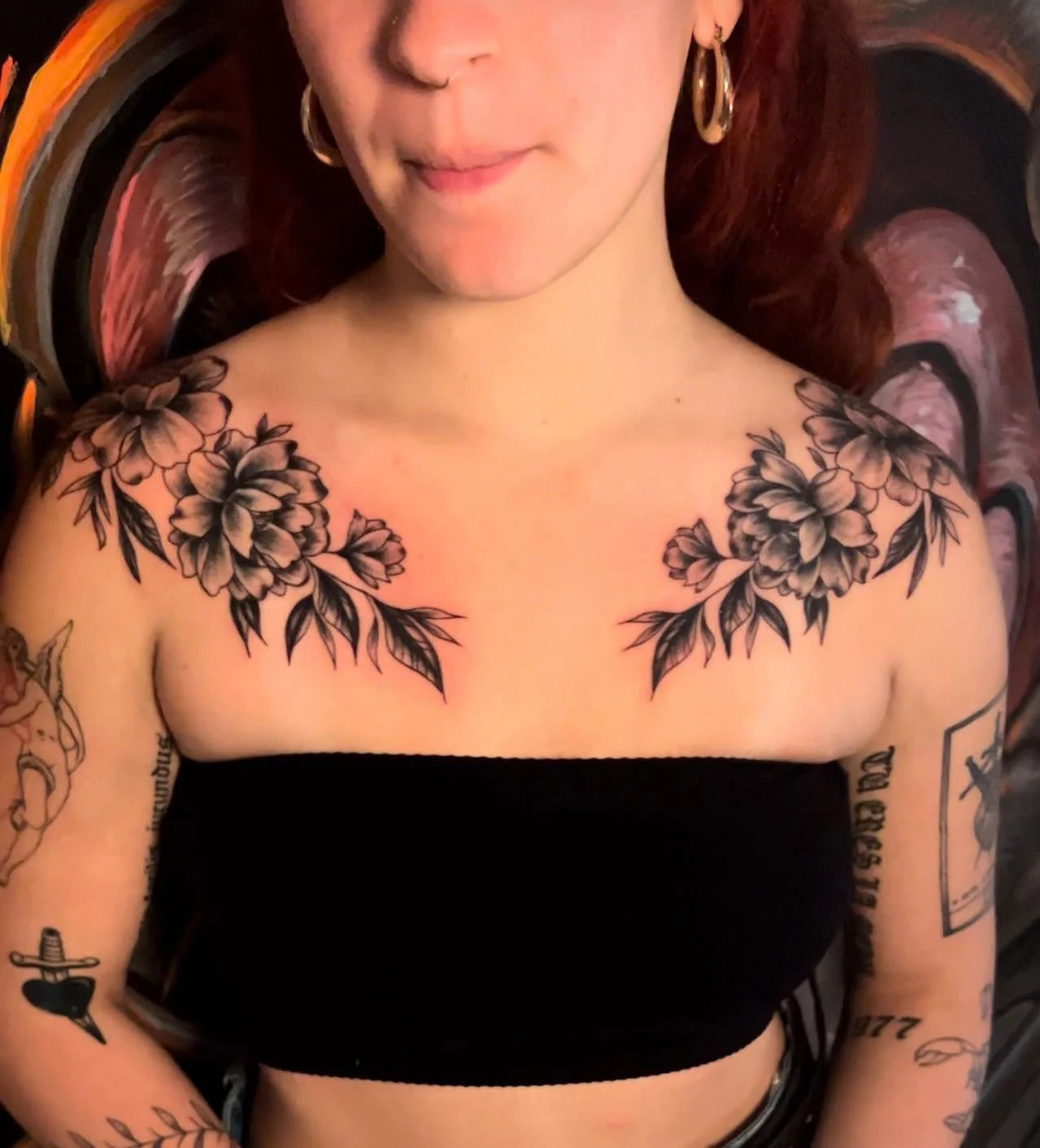 A woman with shoulder-length red hair and earrings displays a large floral tattoo across her upper chest and shoulders. She is wearing a black tube top, and her arms have multiple tattoos, including a black rose and dagger on her left arm. The backgr