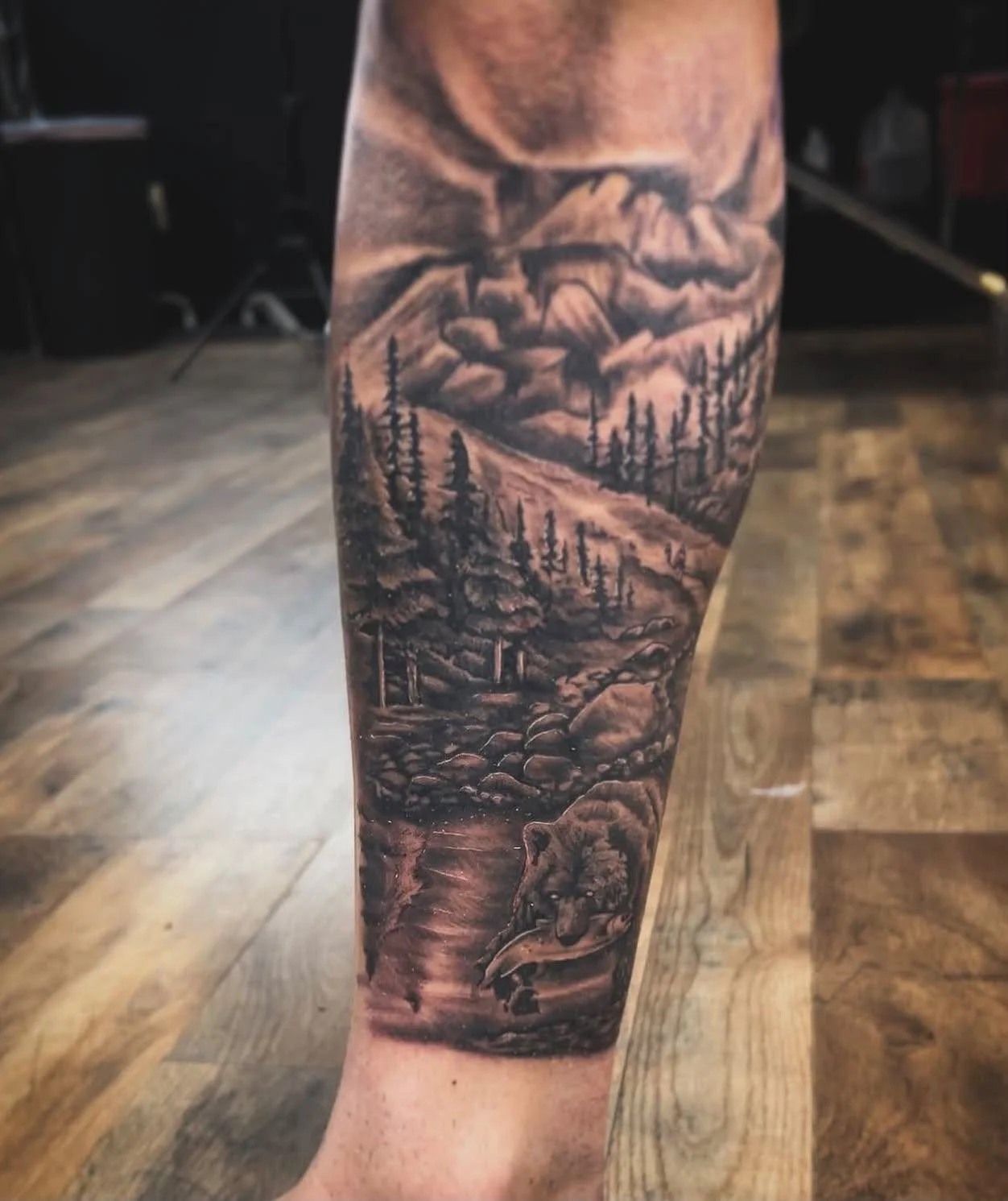 Tattoo of a mountain landscape with trees and a bear on a person's arm.
