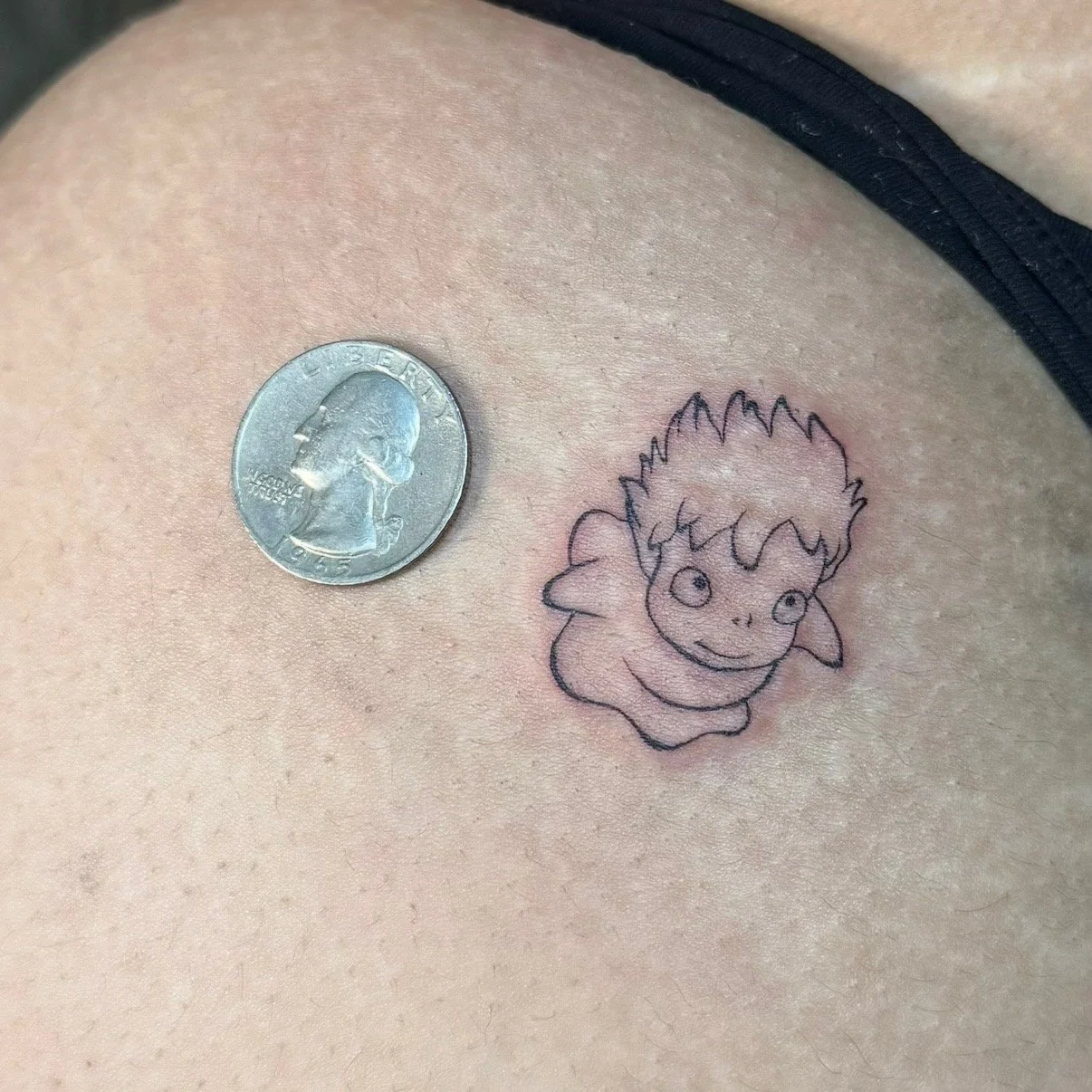 Close-up of a person's skin with a black ink tattoo of a cartoonish, smiling creature with large eyes and spiky hair, next to a U.S. quarter to show scale.