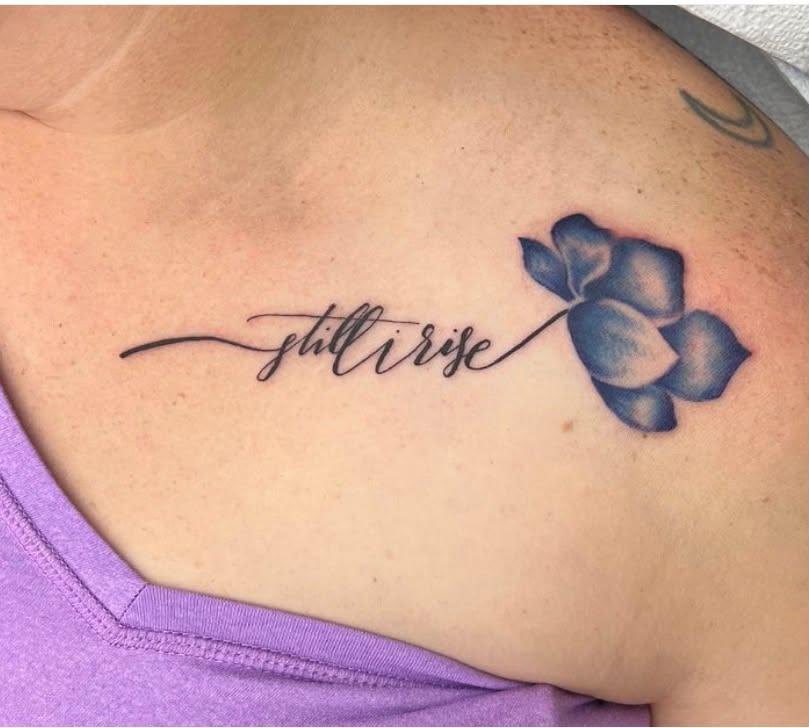 Tattoo of the words 'still rise' in cursive script along with a shaded blue flower on a person's shoulder.