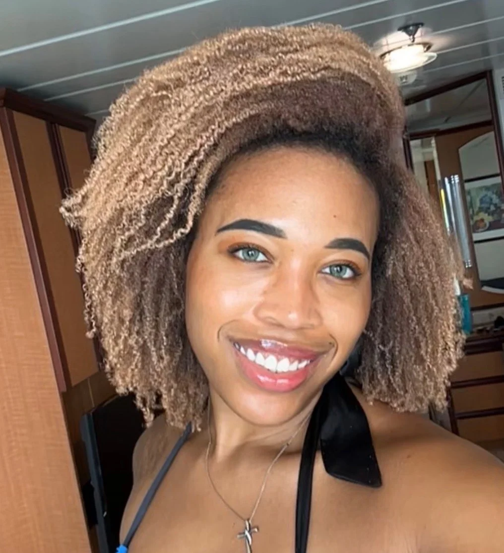 A woman with light brown curly hair, blue eyes, and a bright smile, taking a selfie indoors.