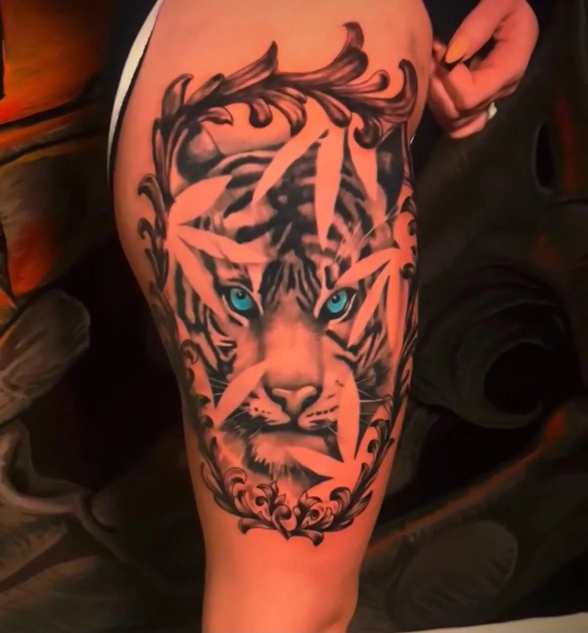 Tattoo of a realistic tiger's face with blue eyes, surrounded by decorative floral and leaf elements, on a person's arm.