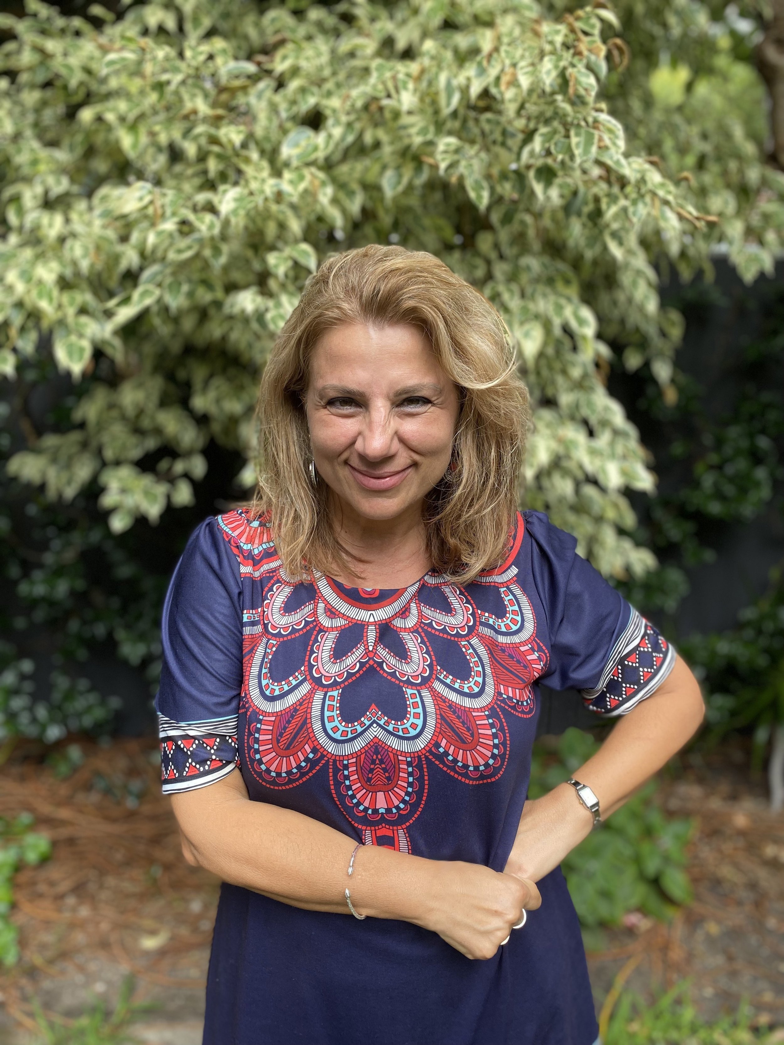 Woman in blue dress with red and white patterns, standing in front of green foliage.
