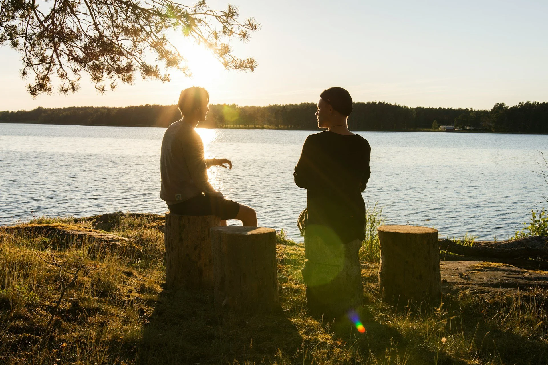 Two people sitting on wooden logs near a lake during sunset, with silhouettes against the water and trees in the background.
