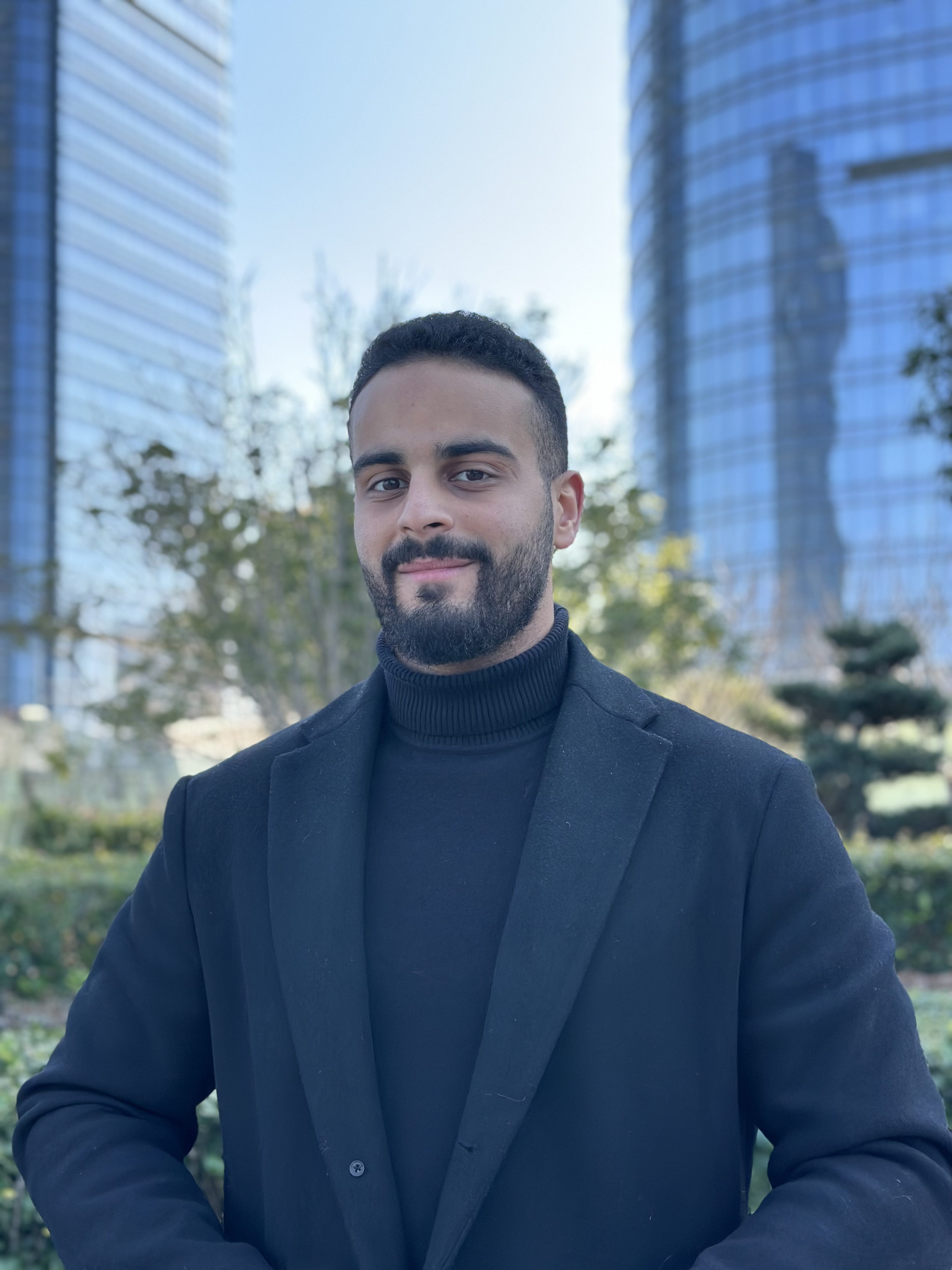 A man with dark hair and a beard, wearing a black turtleneck and blazer, standing outdoors with modern glass buildings and trees in the background.