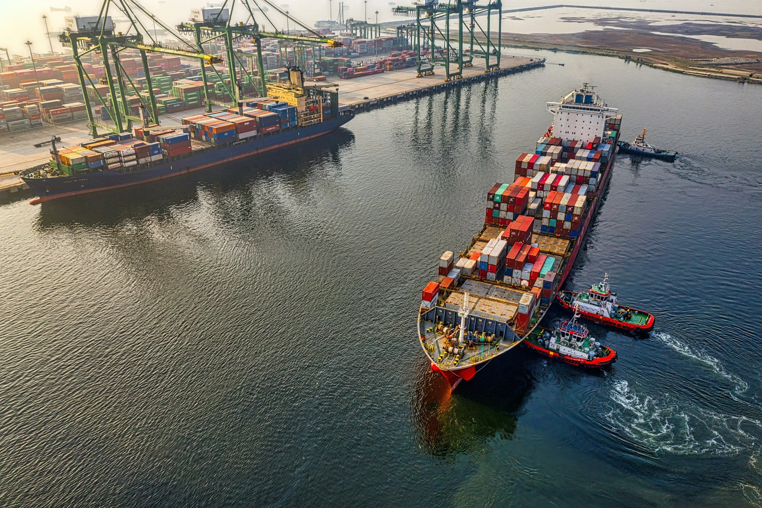 An aerial view of cargo ships loaded with containers in a busy port with cranes, and tugboats guiding the ships through the water.