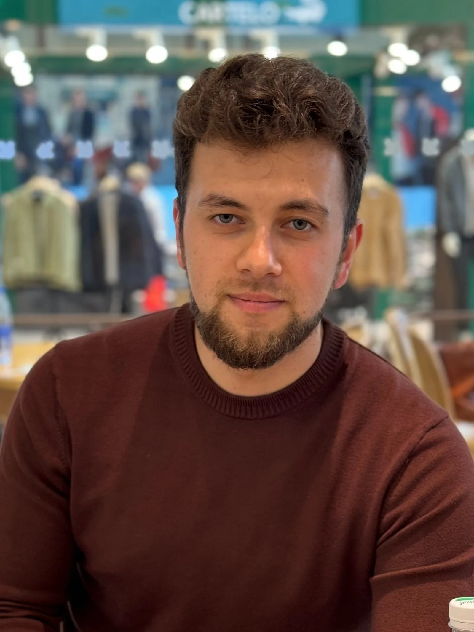 A young man with brown hair and a beard wearing a burgundy shirt, sitting indoors with a blurred background of a clothing store or market.