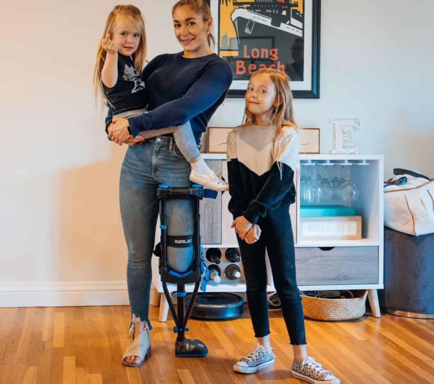 A woman with two young girls standing in a living room with a framed art piece titled 'Long Beach' on the wall behind them. One girl is sitting on a mobility scooter, and the other girl is standing next to them with her hands clasped.