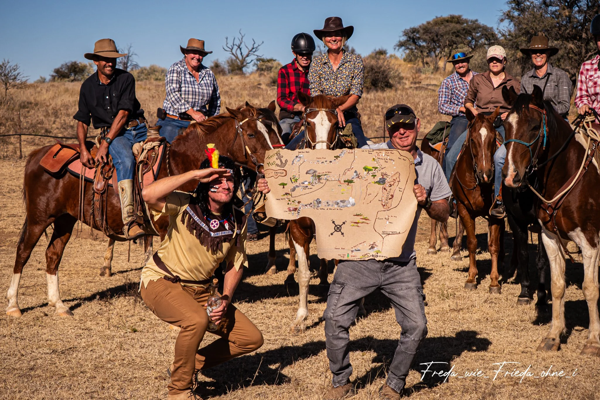 Eine Gruppe von Menschen reitet auf Pferden durch eine trockene, hügelige Landschaft mit einigen Bäumen. Eine Person hält eine Karte und zeigt sie in Richtung Kamera, während die anderen lächelnd in die Kamera schauen. Erlebe Namibia vom Pferderücken