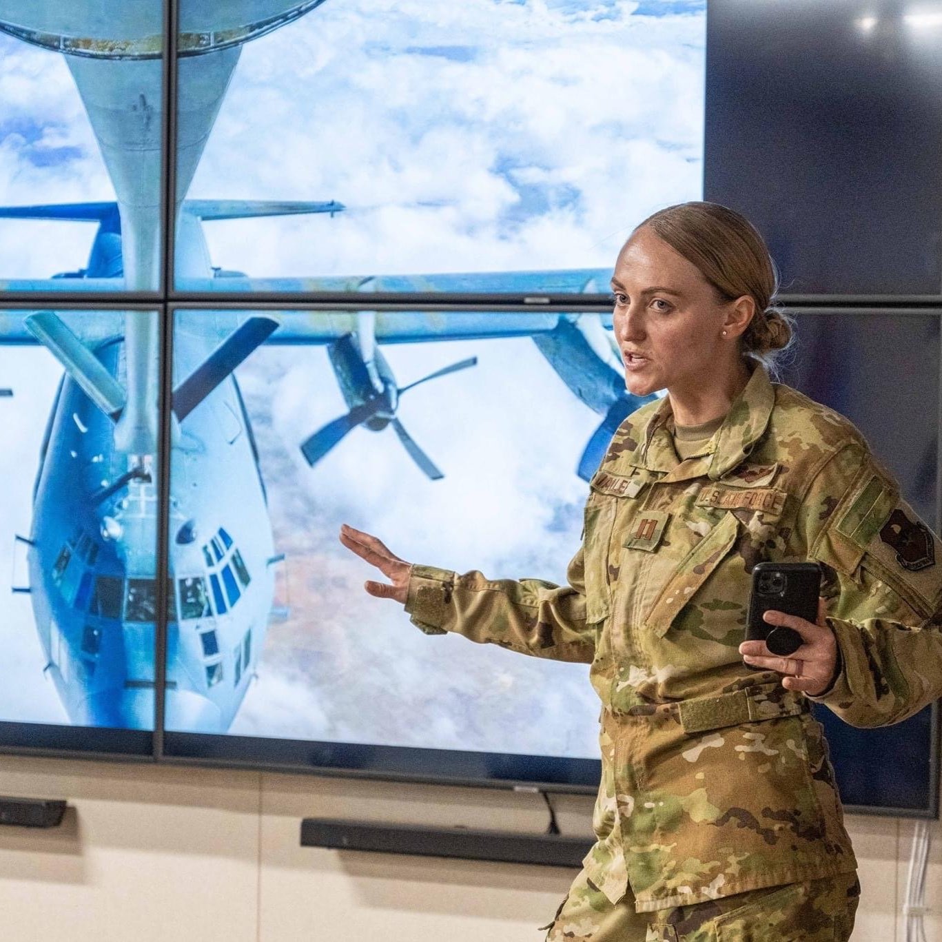A person in camouflage military uniform standing in front of a screen displaying a large aircraft, speaking or presenting.