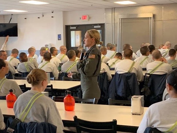 A group of personnel in a dining hall, many wearing gray shirts with reflective suspenders, listen to a woman in a military uniform who is holding a microphone. Red water bottles and jackets are on the tables.