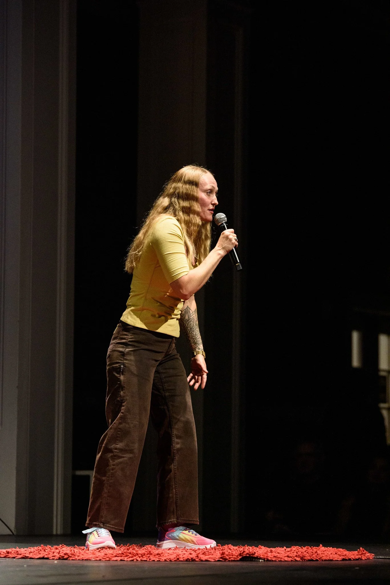 A woman with long curly blonde hair standing on a stage, holding a microphone in her right hand, speaking. She is wearing a yellow top, brown pants, and colorful sneakers. She is standing on a red rug against a dark background.