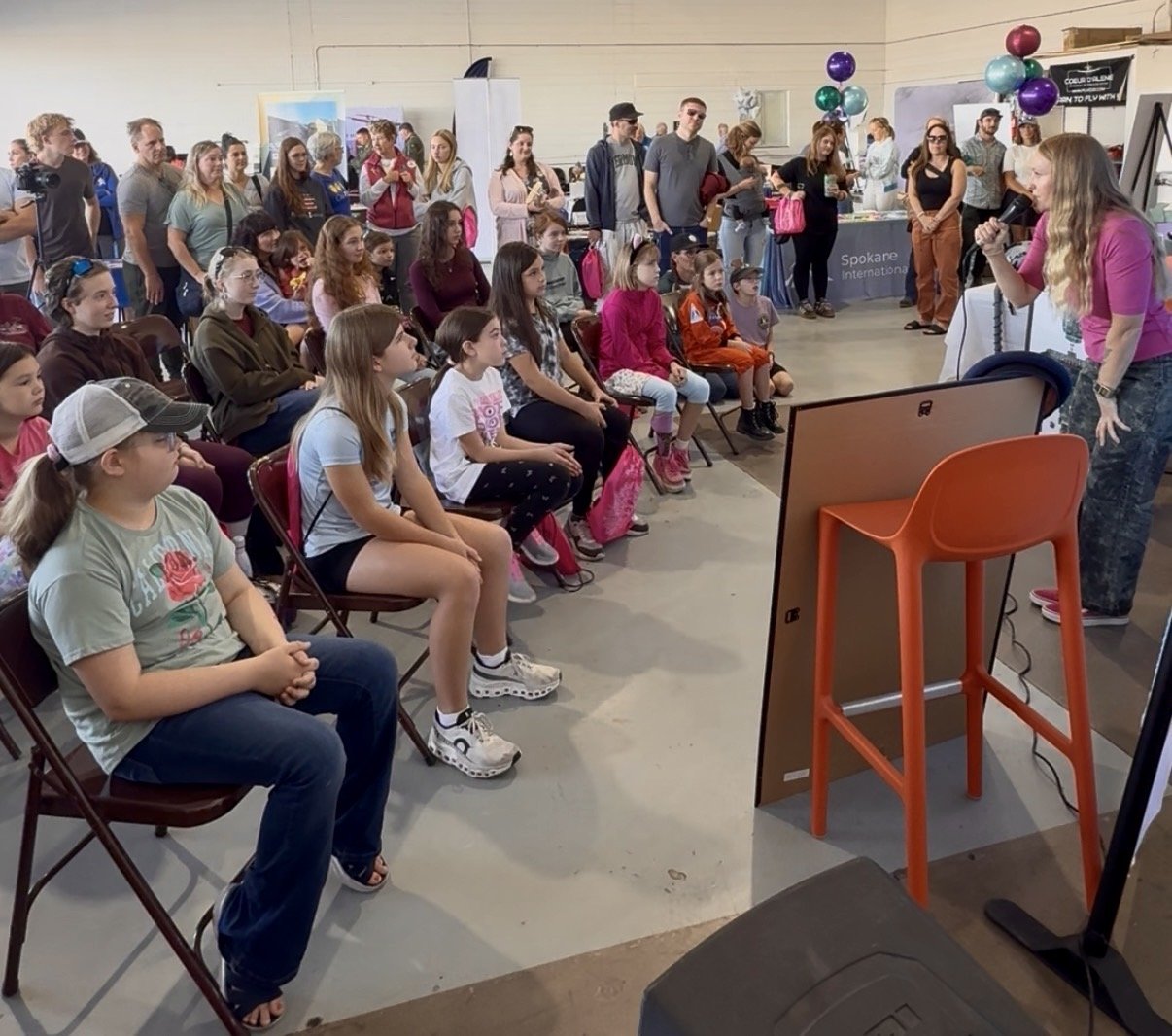 A woman in a pink shirt is speaking or singing into a microphone in front of a group of children seated on chairs, attending an indoor event. Several adults are standing in the background, and there are colorful balloons and display tables around the