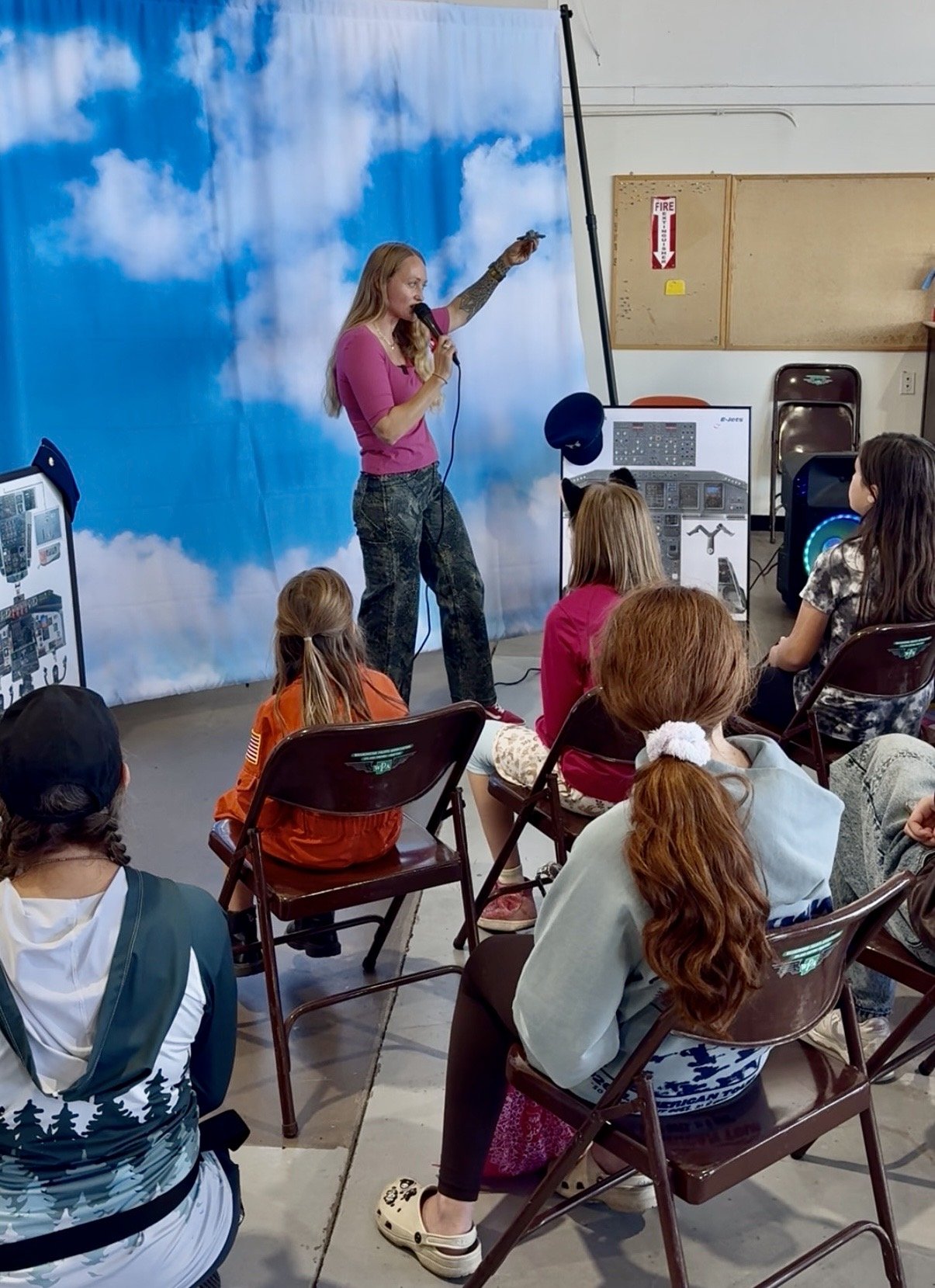 A woman with long blonde hair, wearing a pink top and camouflage pants, is standing on a stage-like area in front of a blue sky cloud backdrop, holding a microphone and gesturing with her hand. She is speaking to a seated group of children and teenag
