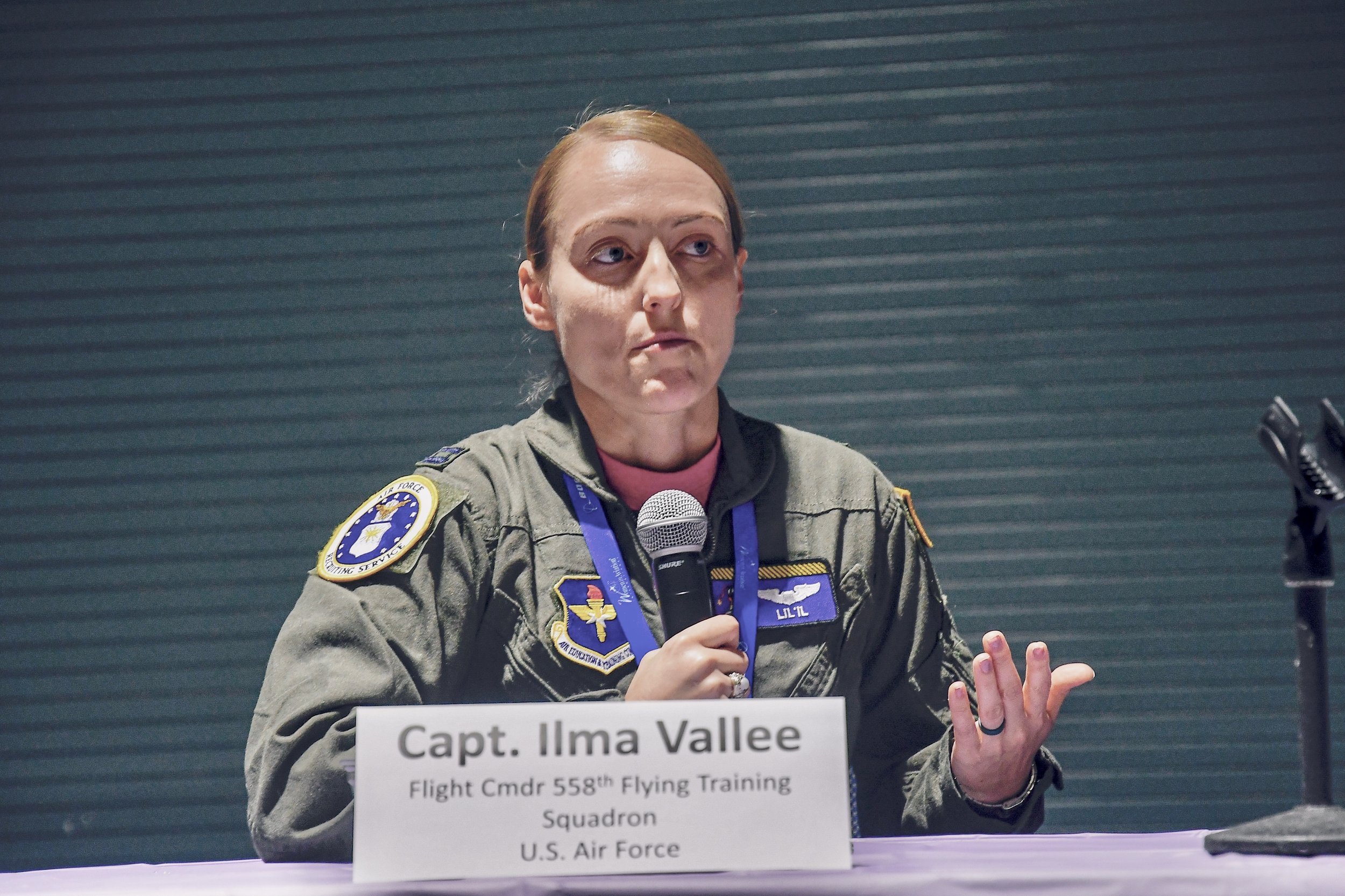 U.S. Air Force officer in uniform speaking at a press conference, holding a microphone in her right hand. The nameplate in front of her reads "Capt. Ilma Vallee, Flight Cmdr 558th Flying Training Squadron, U.S. Air Force."