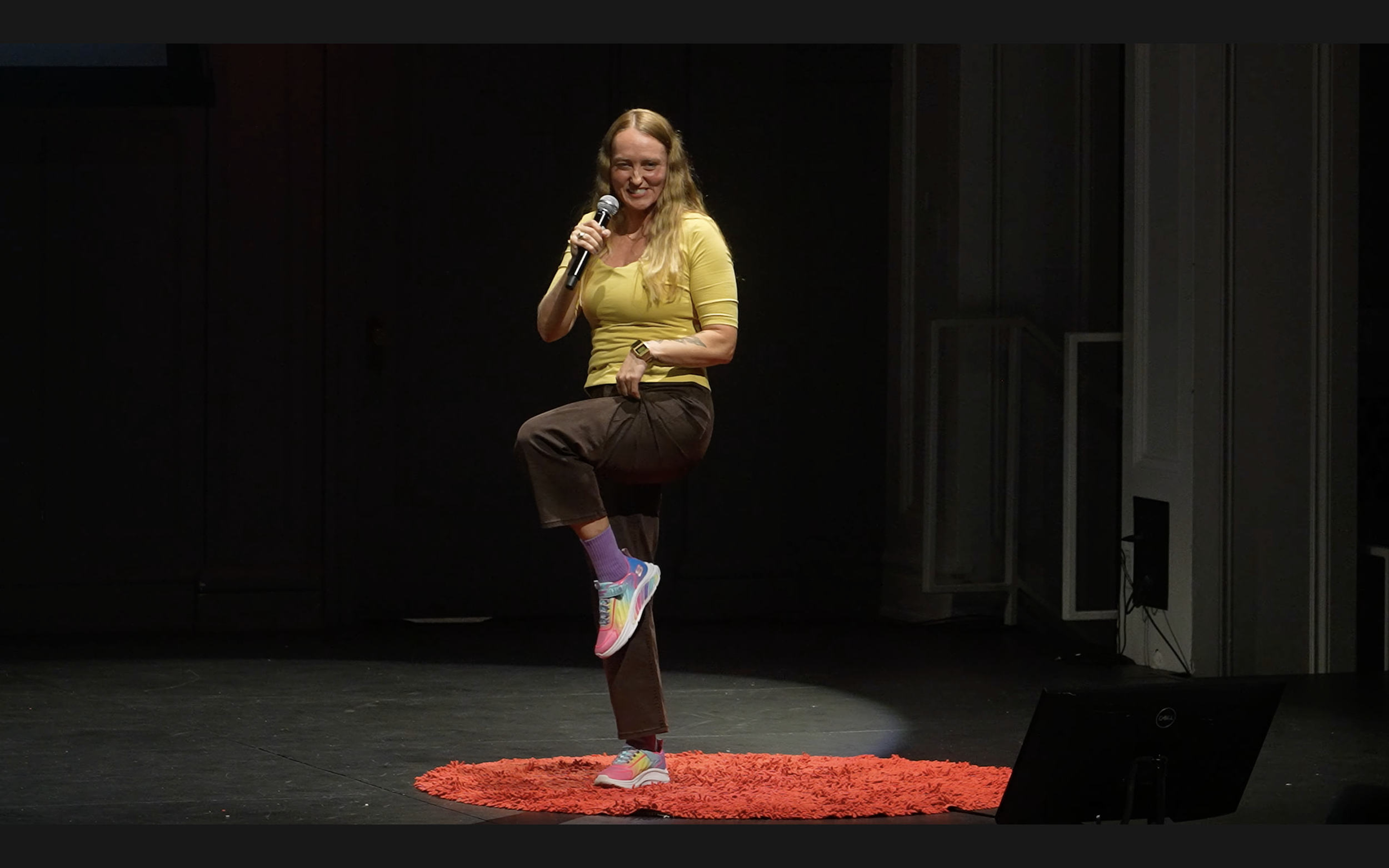 A woman with long blonde hair, wearing a yellow top, brown pants, colorful sneakers, and purple socks, sits on a small stool on a stage with a red rug, holding a microphone and smiling.
