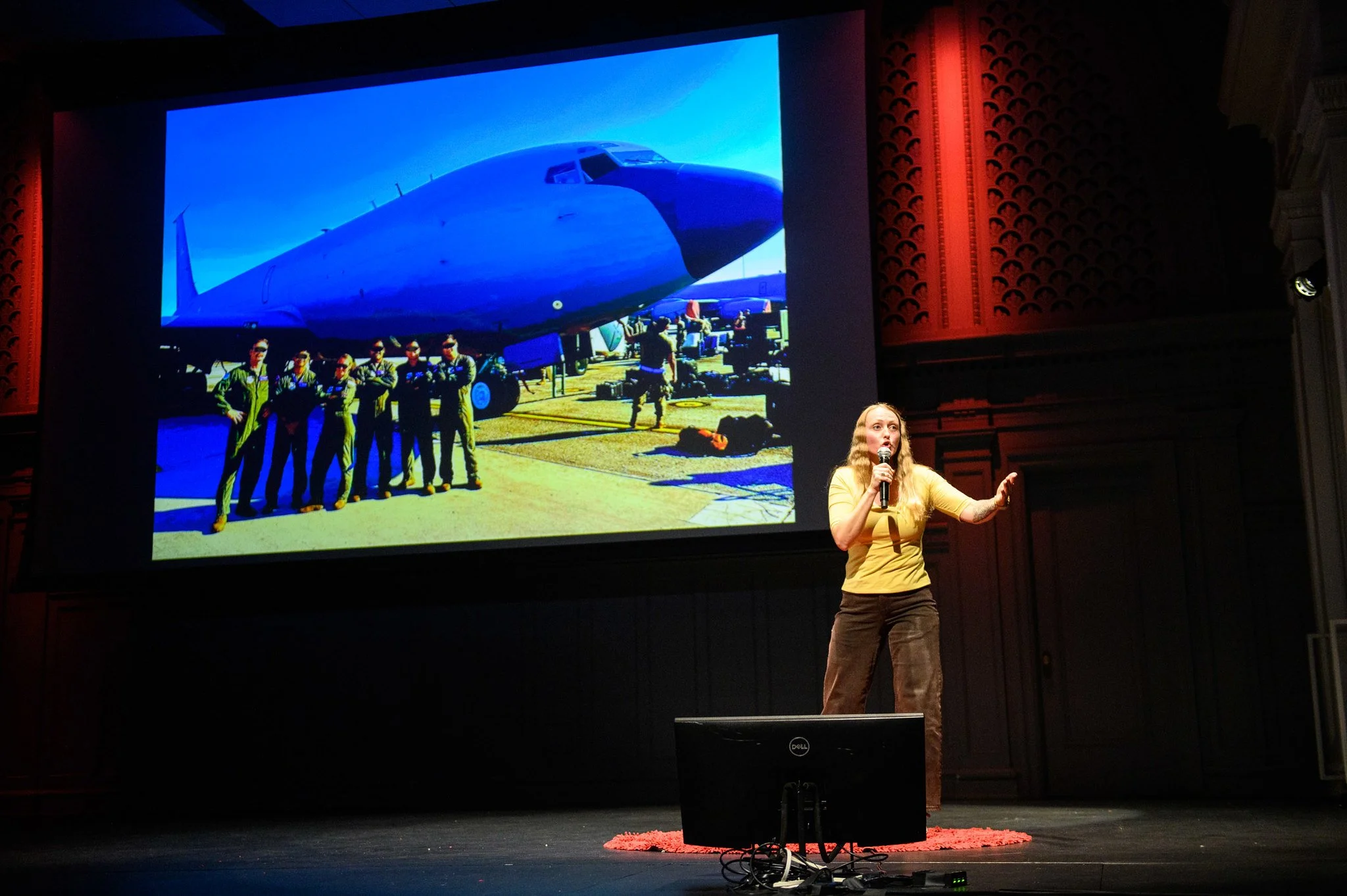 A woman in a yellow top giving a presentation on stage with a microphone, in front of a large screen displaying a photo of a military aircraft and crew members.