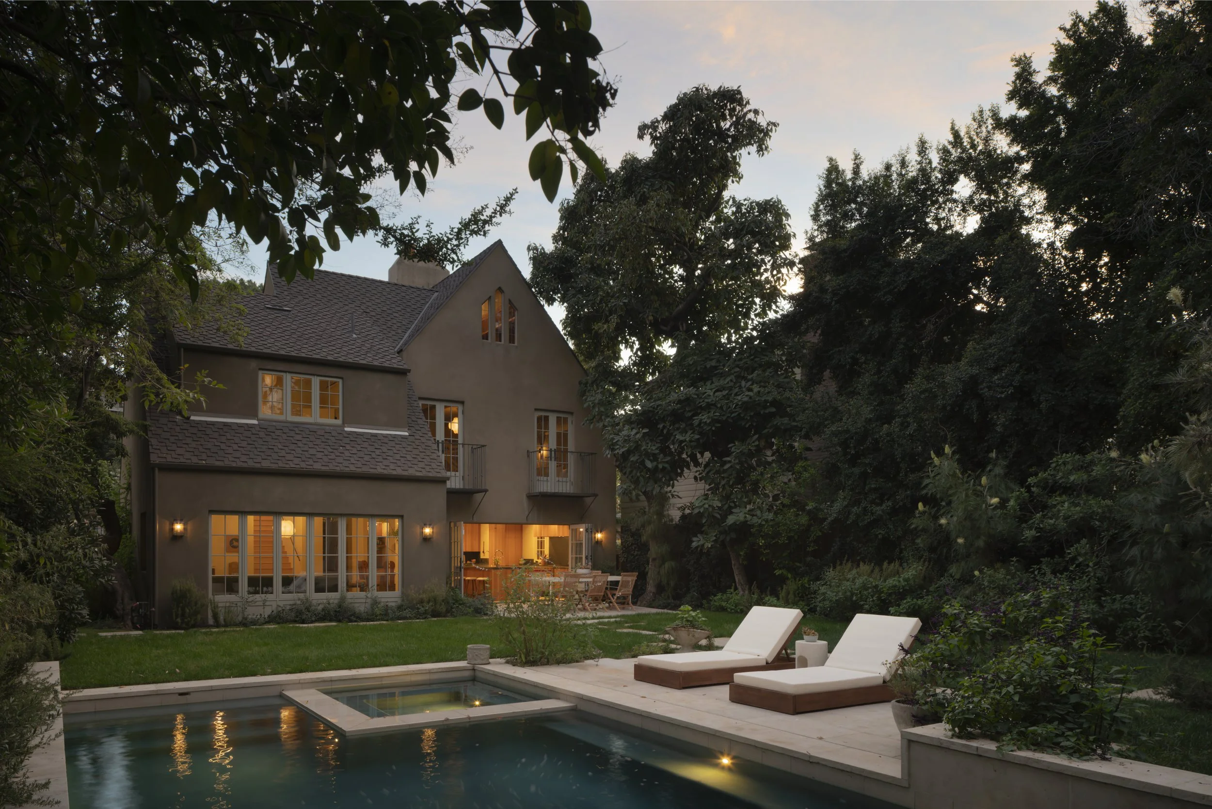 A backyard view of a modern house with lit windows, a swimming pool, and two lounge chairs with cushions at dusk, surrounded by lush trees and greenery.