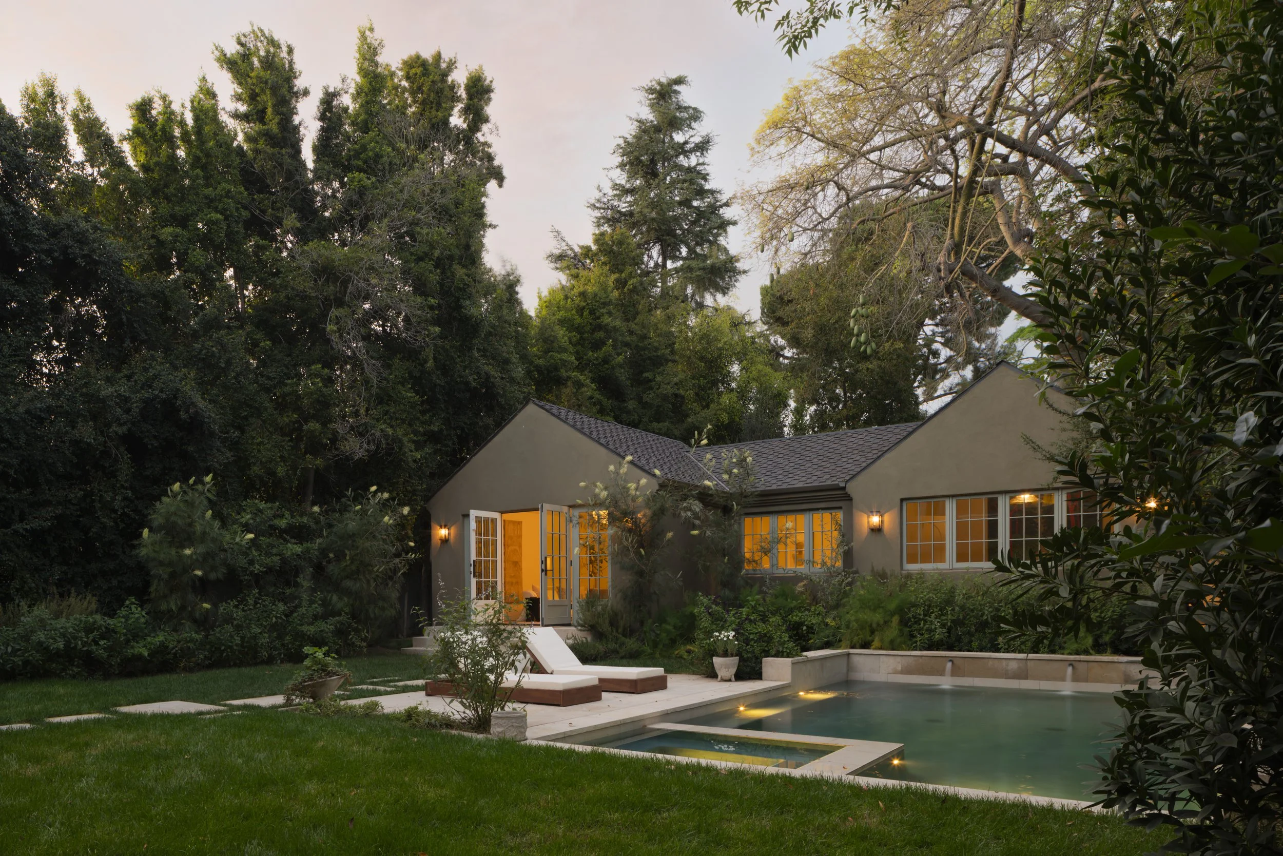 A backyard with a swimming pool, lounge chairs, and a house with illuminated windows at dusk, surrounded by trees and greenery.