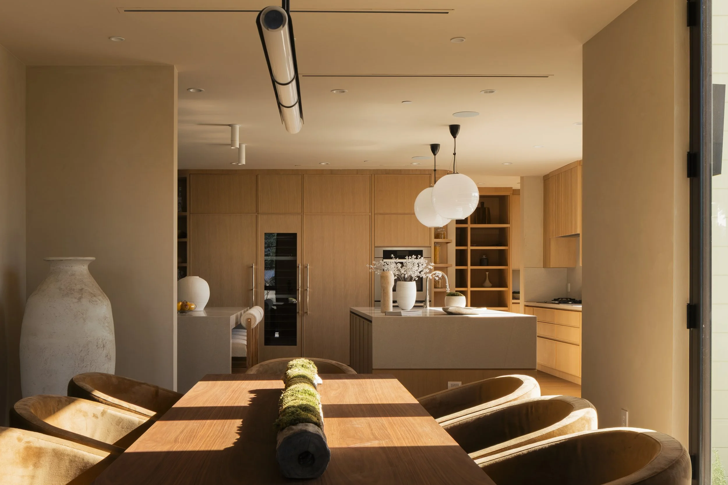 Modern kitchen and dining area with wooden cabinets, an island, pendant lights, and a dining table with chairs, illuminated by natural sunlight.