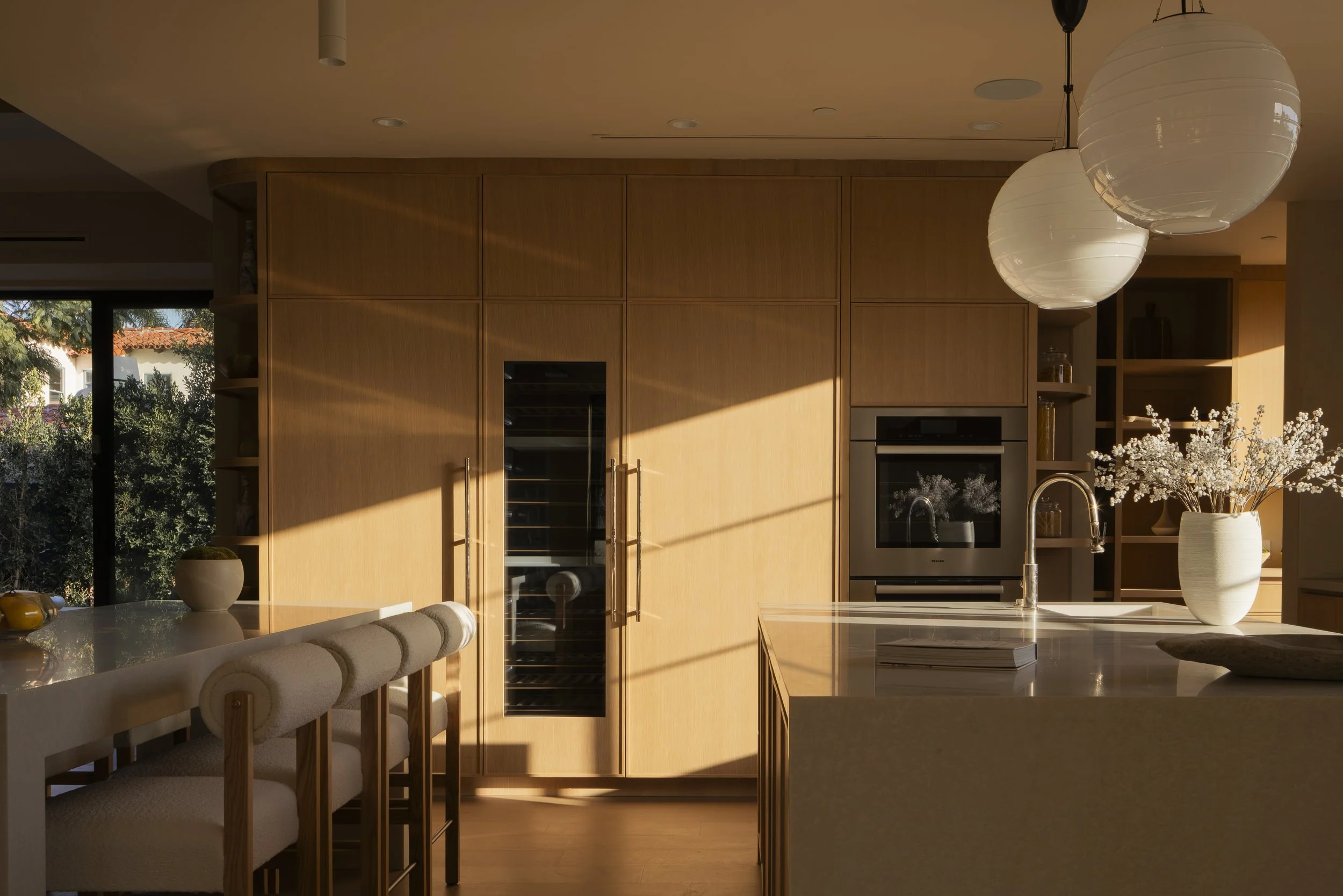 A modern kitchen with wooden cabinets, an oven, a wine fridge, and a large kitchen island with a sink. Sunlight streams through a window, casting shadows on the cabinets. There are two hanging white paper lanterns and a vase with white flowers on the