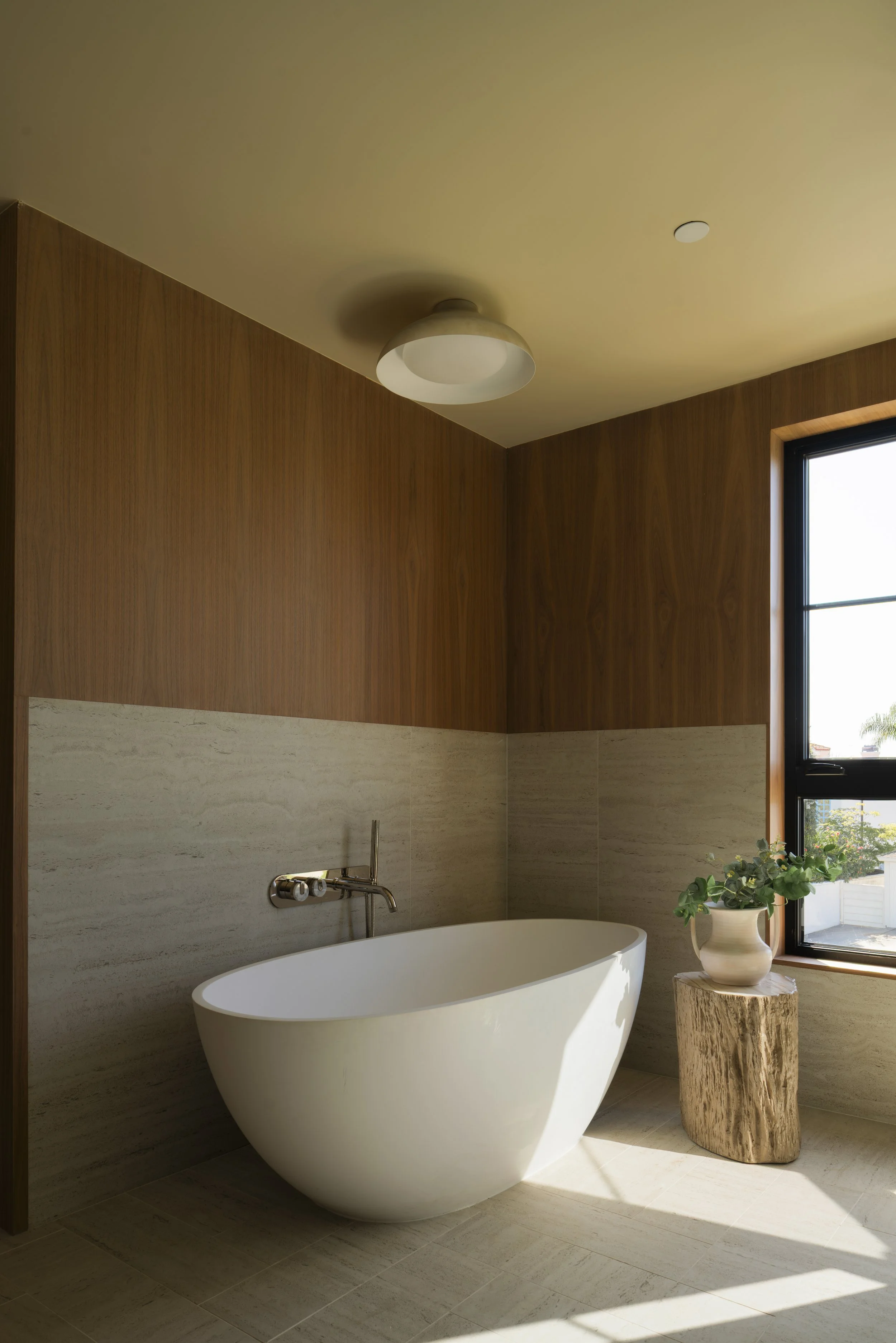 Modern bathroom with a white freestanding bathtub, a wooden wall, beige tile walls and floor, a round ceiling light, a window with a plant on a wooden stump, and natural sunlight.