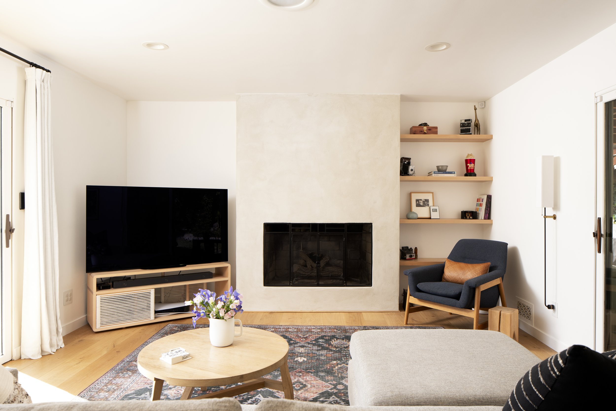 Living room with white walls, fireplace, flat-screen TV on a wooden stand, beige rug, coffee table with vase of flowers, blue armchair with pillow, and floating wooden shelves with books and decor.
